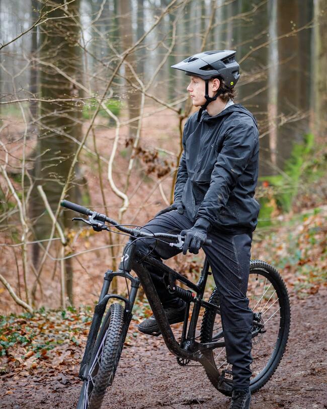 Ein Mountainbiker in schwarzer Sportkleidung, Handschuhen und Helm sitzt auf einem schwarzen Mountainbike und schaut im Profil nach links. Er befindet sich auf einem erdigen Pfad in einem herbstlichen Wald mit kahlen Bäumen und Laub am Boden.