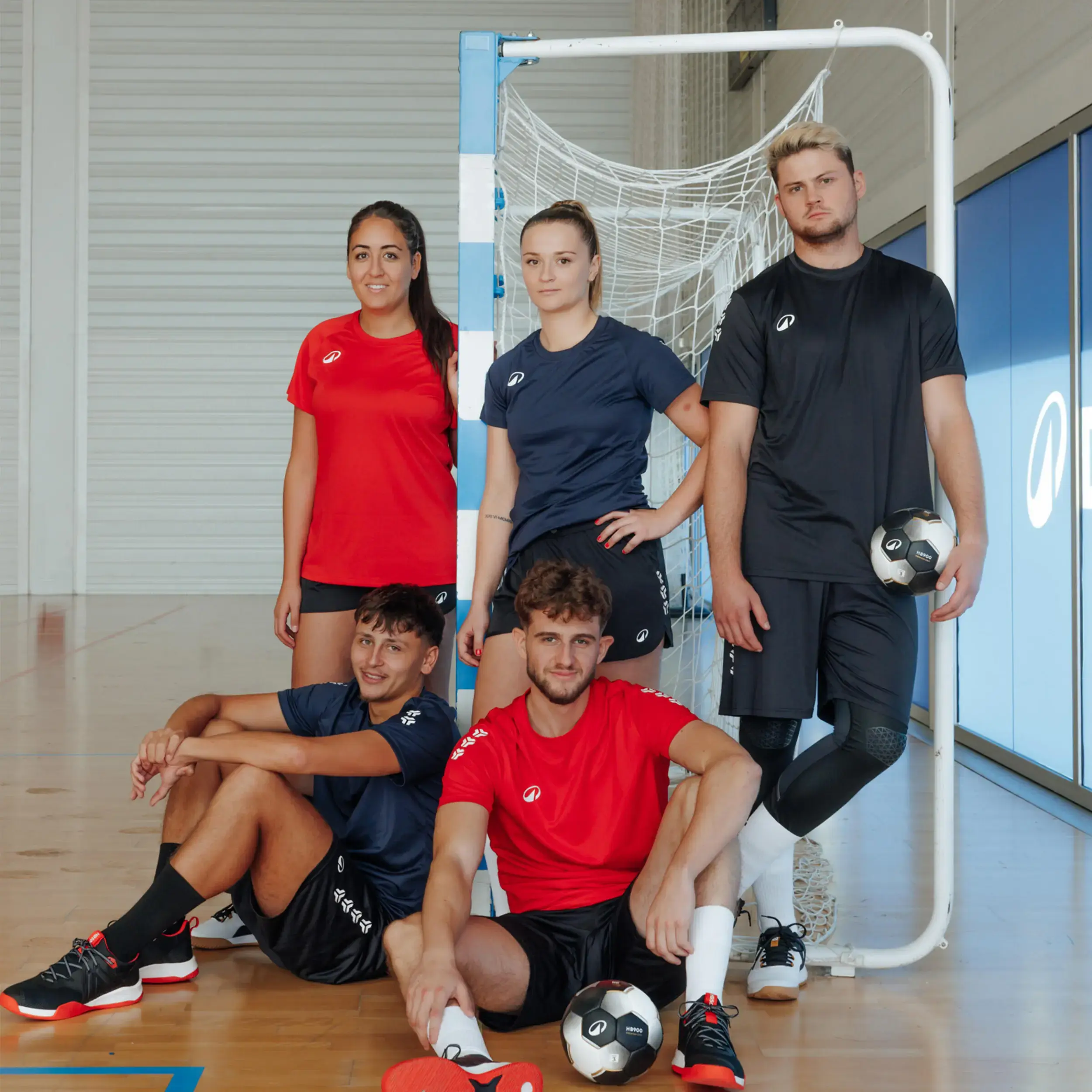 Eine Gruppe von fünf Handballer:innen in roter und schwarzer Kipsta-Kleidung posiert mit einem Kipsta-Ball vor einem Hallentor.