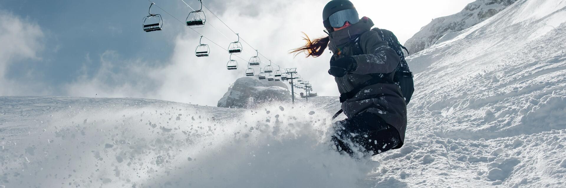 Kobieta w kasku i w goglach jeżdżąca na używanej desce snowboardowej w tle orczyk