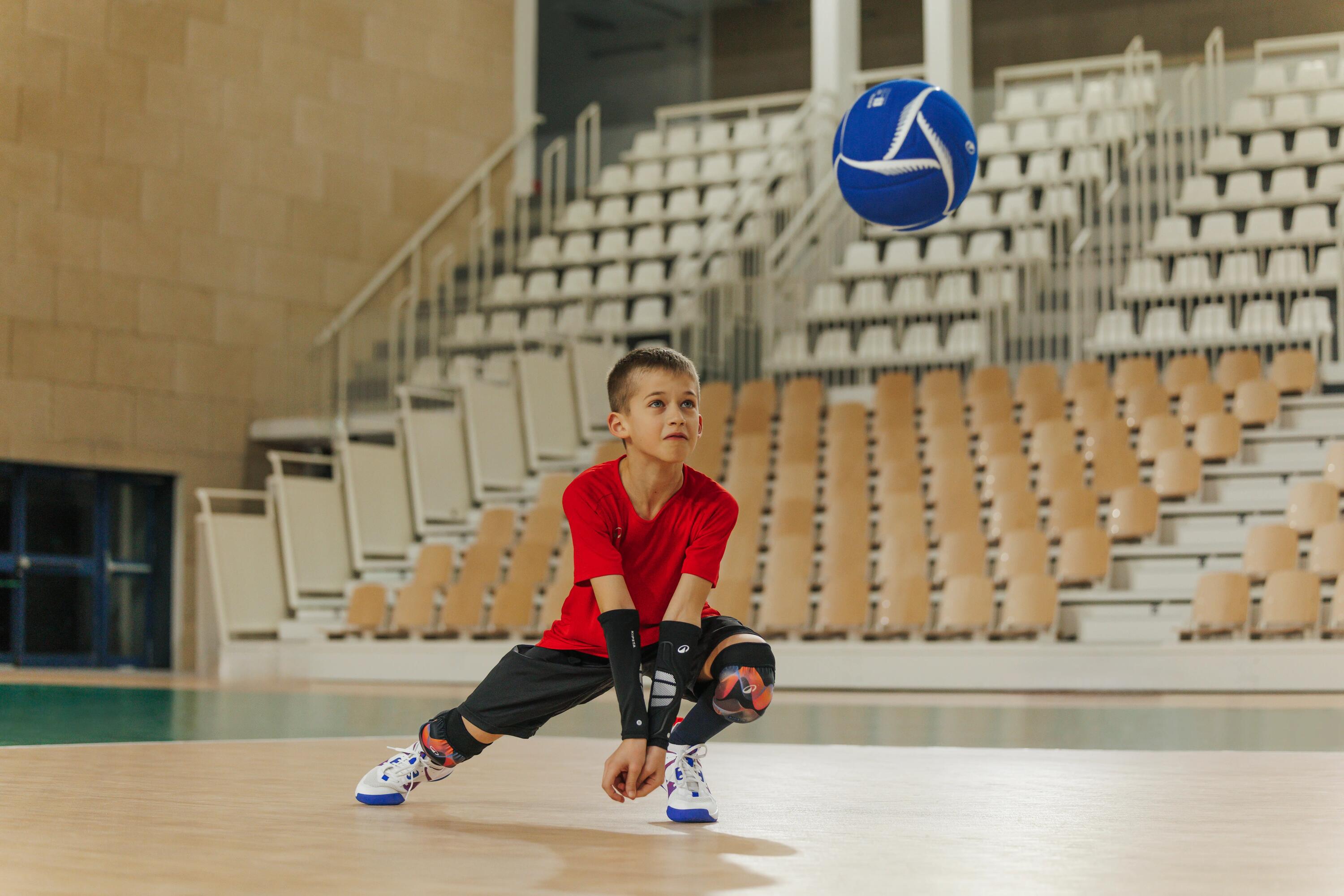 Ein Jungein rotem Shirt und schwarzer Sportkleidung geht in einer großen Halle tief in die Hocke, um einen blau-weißen Volleyball zu pritschen oder zu baggern.