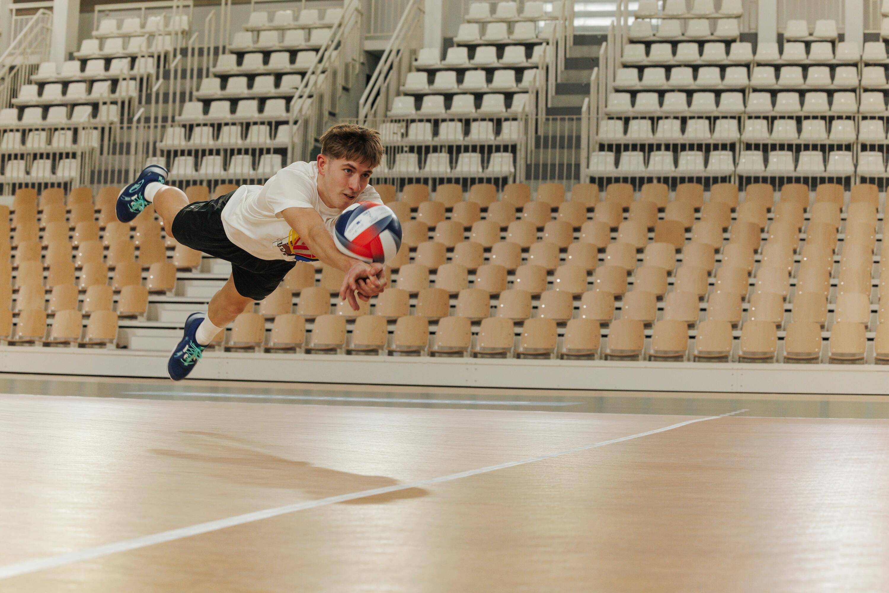 Ein junger Mann springt zu einem Volleyball, um ihn zu baggern. Er befinder sich in einer Halle mit Tribüne.