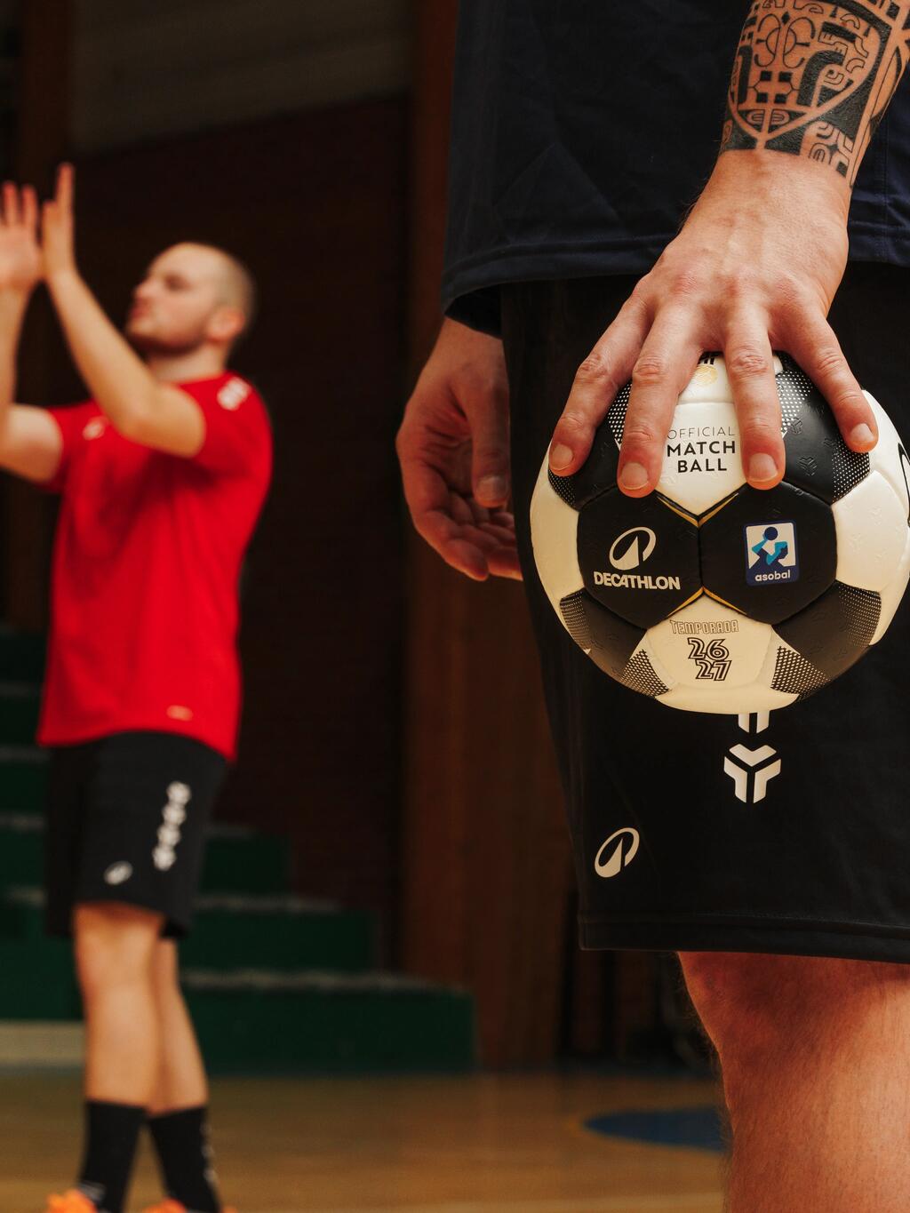 Nahaufnahme in einer Sporthalle: Im Vordergrund hält ein tätowierter Mann einen schwarz-weißen Decathlon-Handball an seiner Hüfte. Im unscharfen Hintergrund wirft ein anderer Spieler in rotem Trikot einen Handball in die Luft.