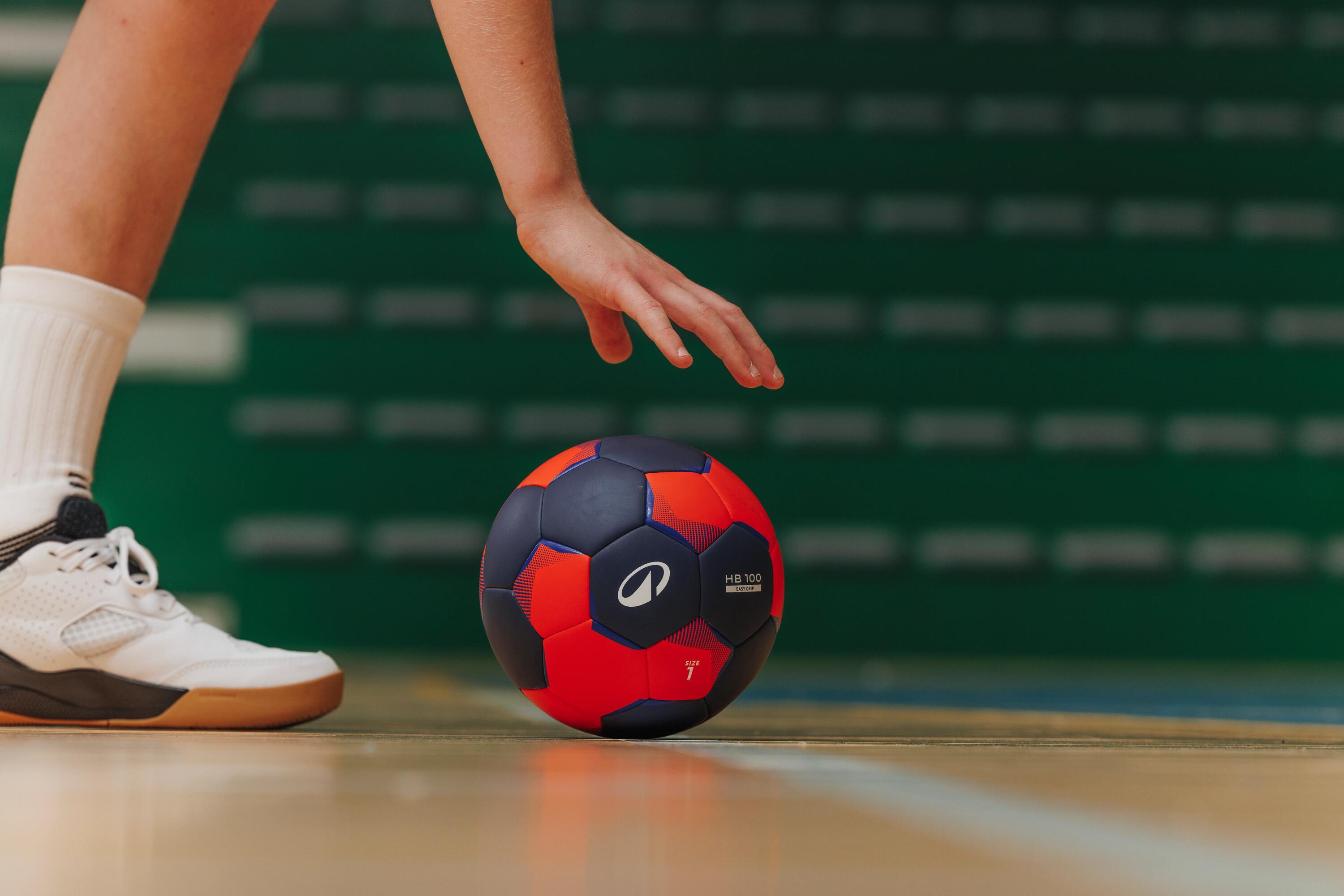 Menschliche Hand greift nach einem Kipsta H100 Ball, der auf einem Hallenboden liegt.