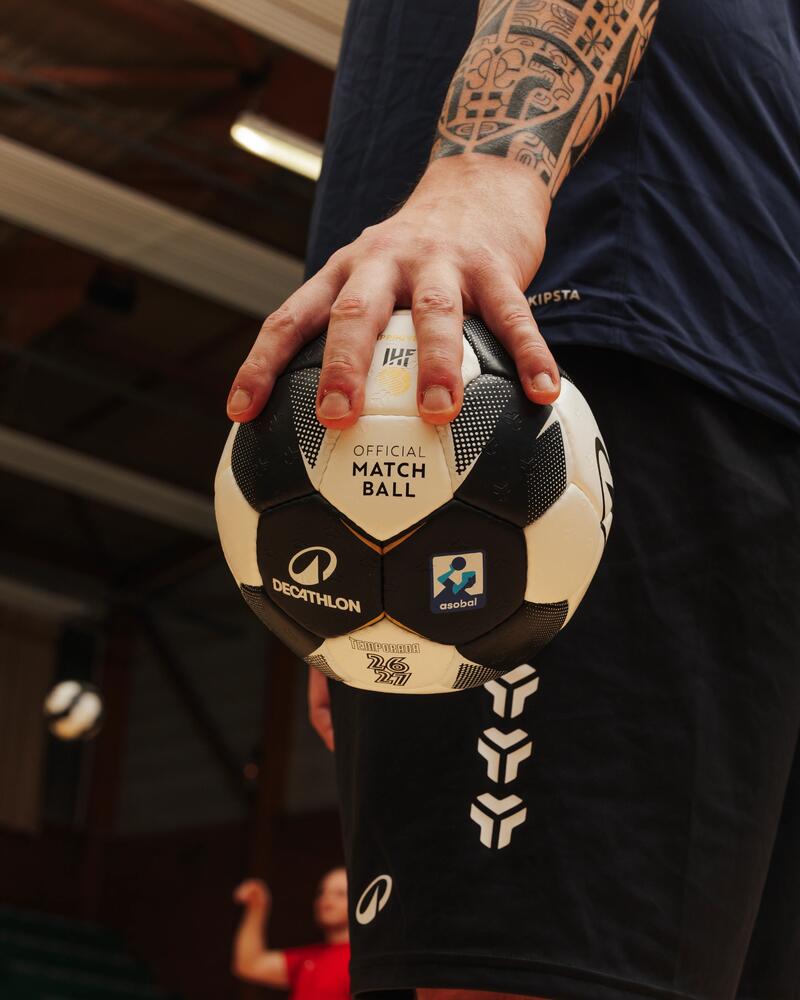 Eine Nahaufnahme zeigt die tätowierte Hand eines Handballspielers, die einen schwarz-weißen Handball in einer Sporthalle hält.