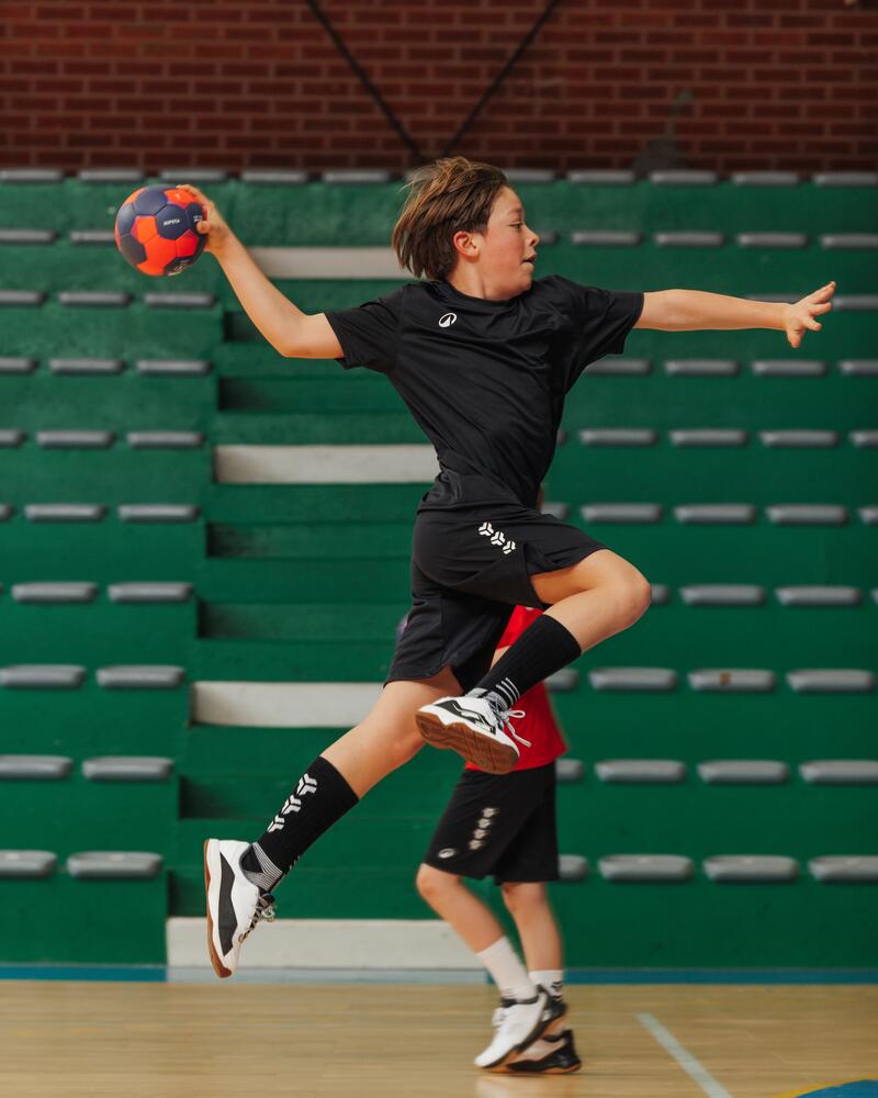Ein junger Handballspieler springt in einer Sporthalle hoch in die Luft, um mit dem Ball in der Hand auf das Tor zu werfen.