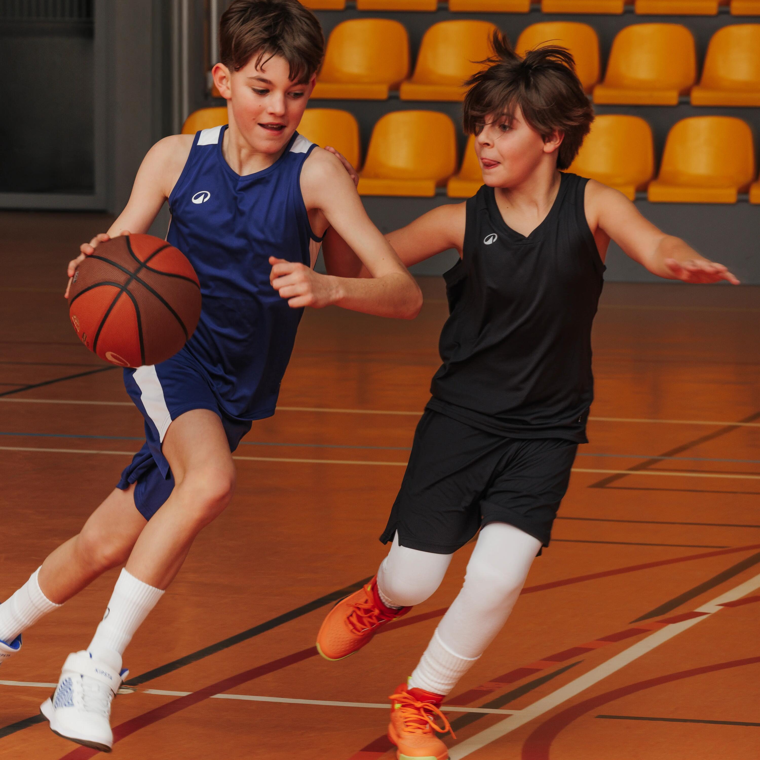 Zwei Jungen spielen auf einem Hallenparkett Basketball, wobei einer im blauen Trikot mit dem Ball dribbelt und der andere im schwarzen Trikot ihn verteidigt.