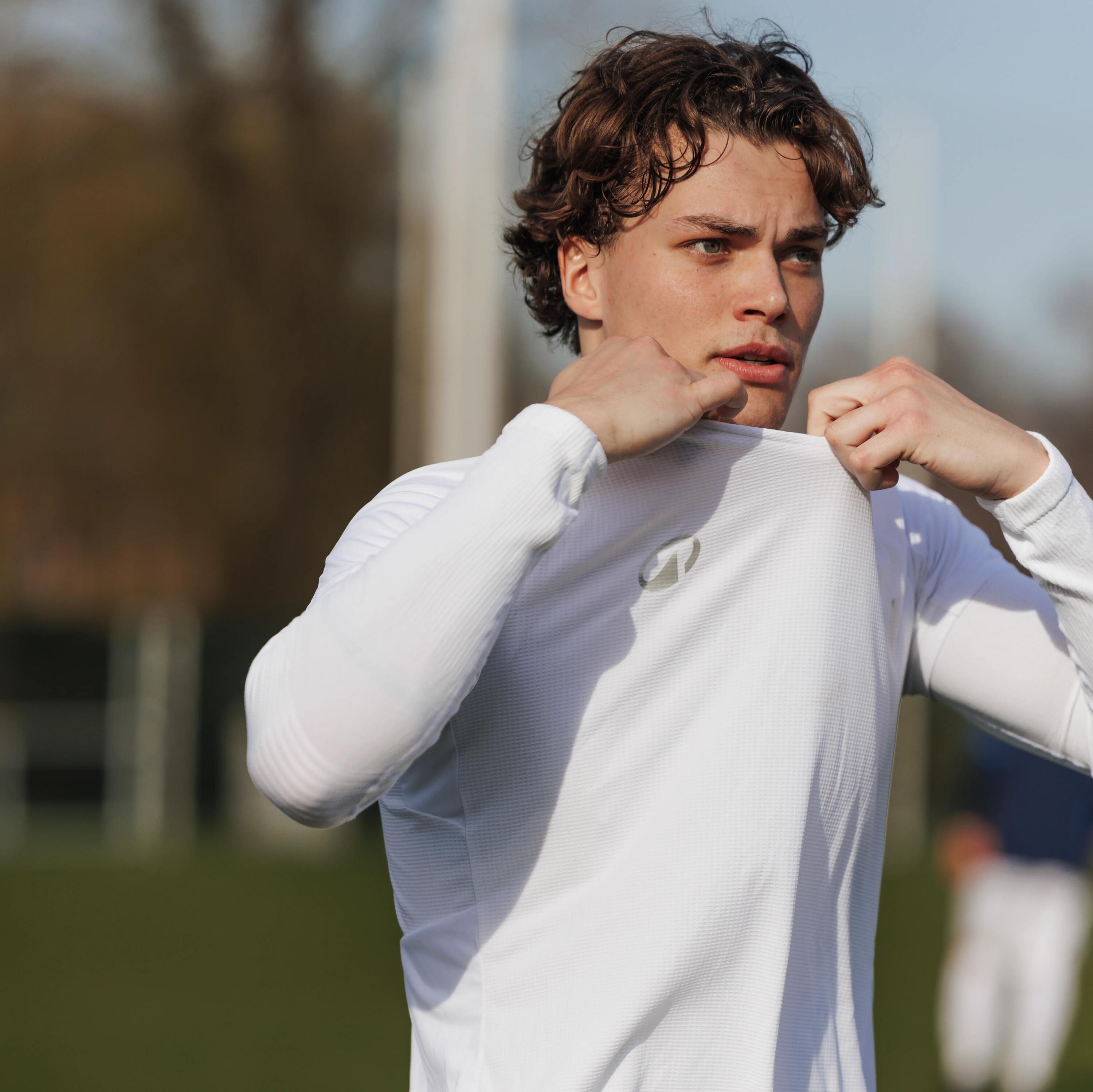 Ein junger, sportlicher Mann mit braunen Haaren steht im warmen Sonnenlicht auf einem Fußballplatz, blickt nach rechts und hält ein blaues Trikot in den Händen.