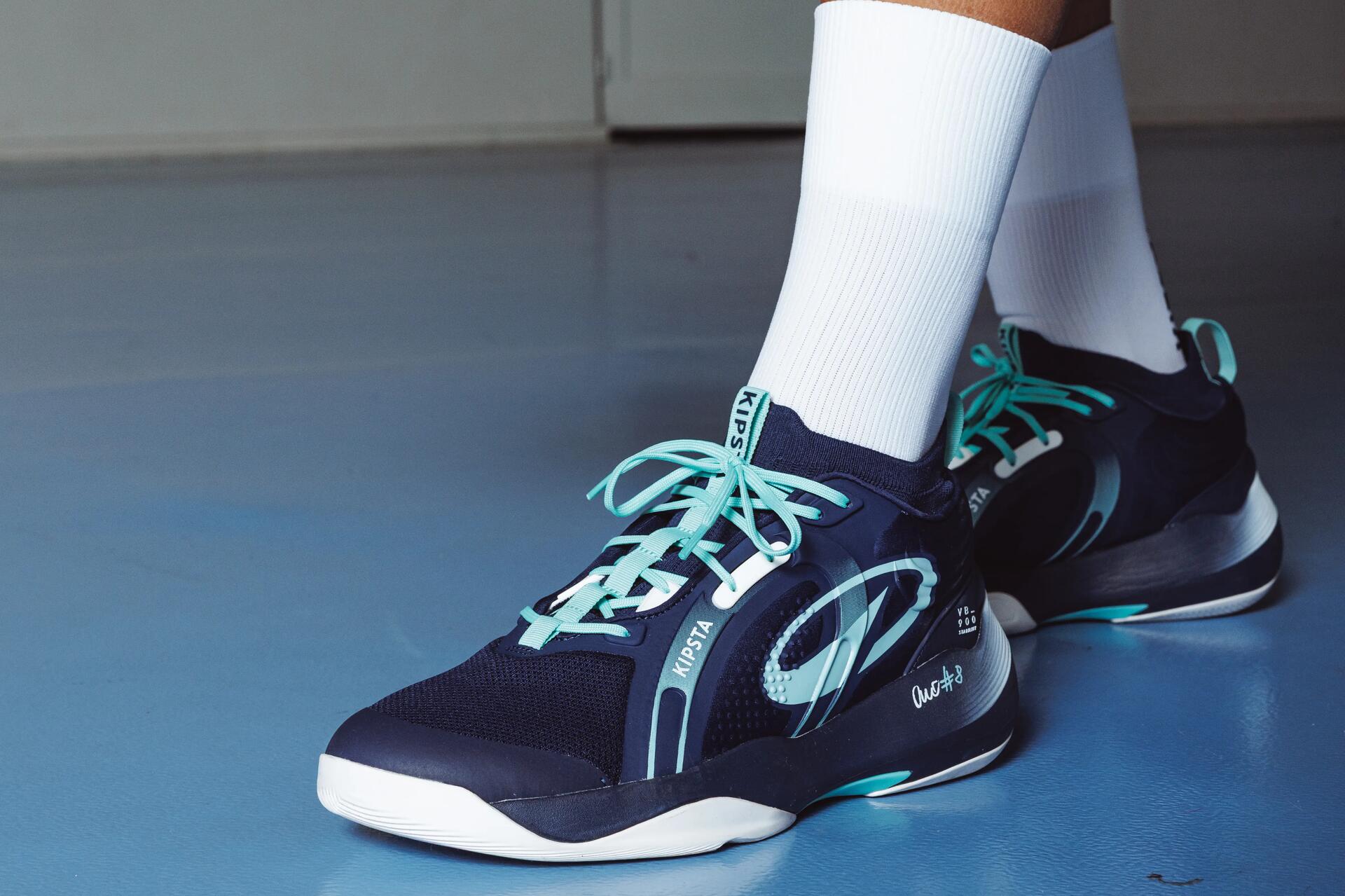 Eine Nahaufnahme zeigt die Beine und Hände einer Volleyballspielerin mit Knieschonern und Sportschuhen, die einen blau-weiß-roten Volleyball hält.