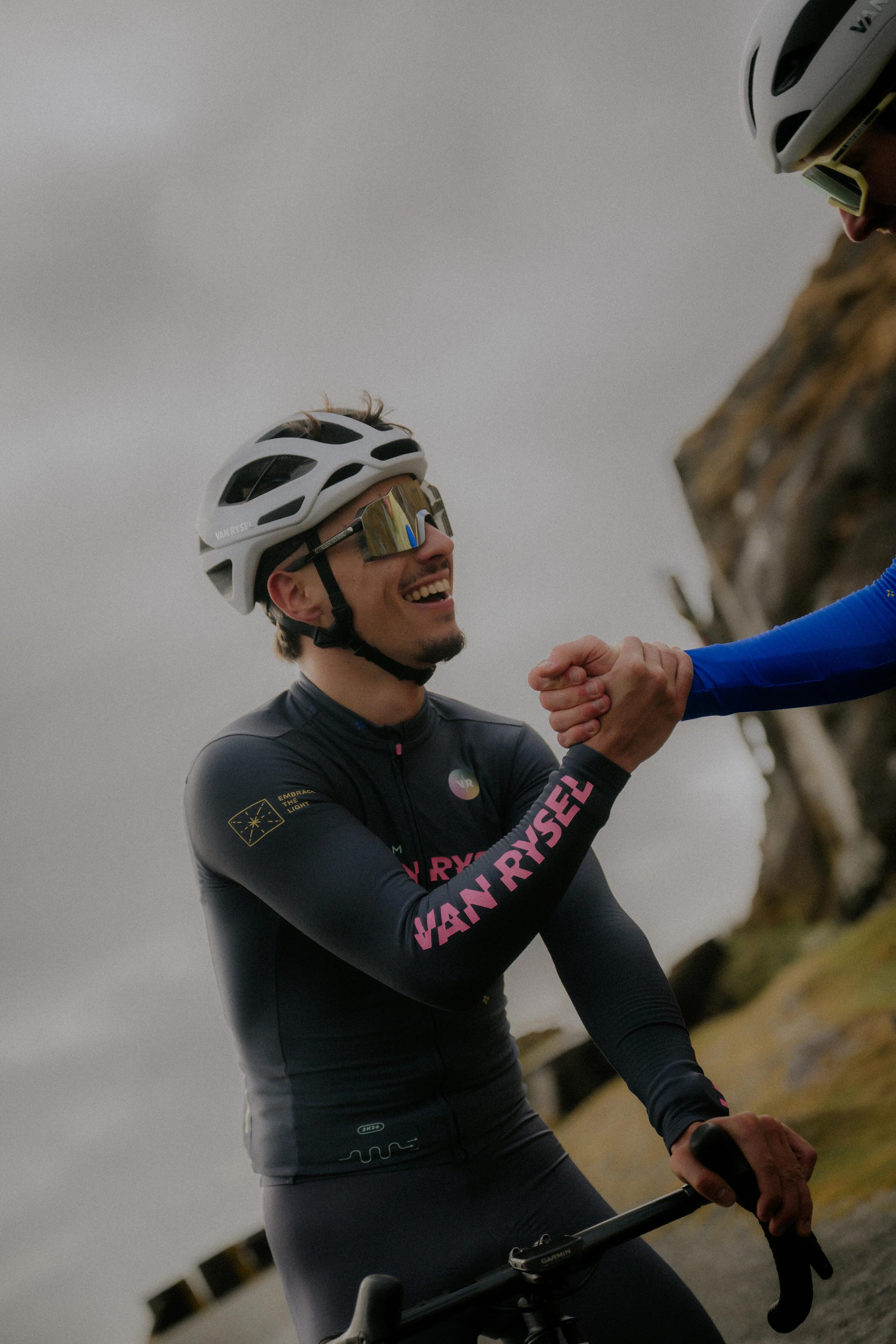 Ein Radrennfahrer in professioneller Decathlon Van Rysel Team-Bekleidung fährt auf einem hellgrünen Rennrad eine leere Landstraße entlang. Er trägt einen hellgrünen Helm, eine große Sportbrille und ein blau-türkises Trikot.
