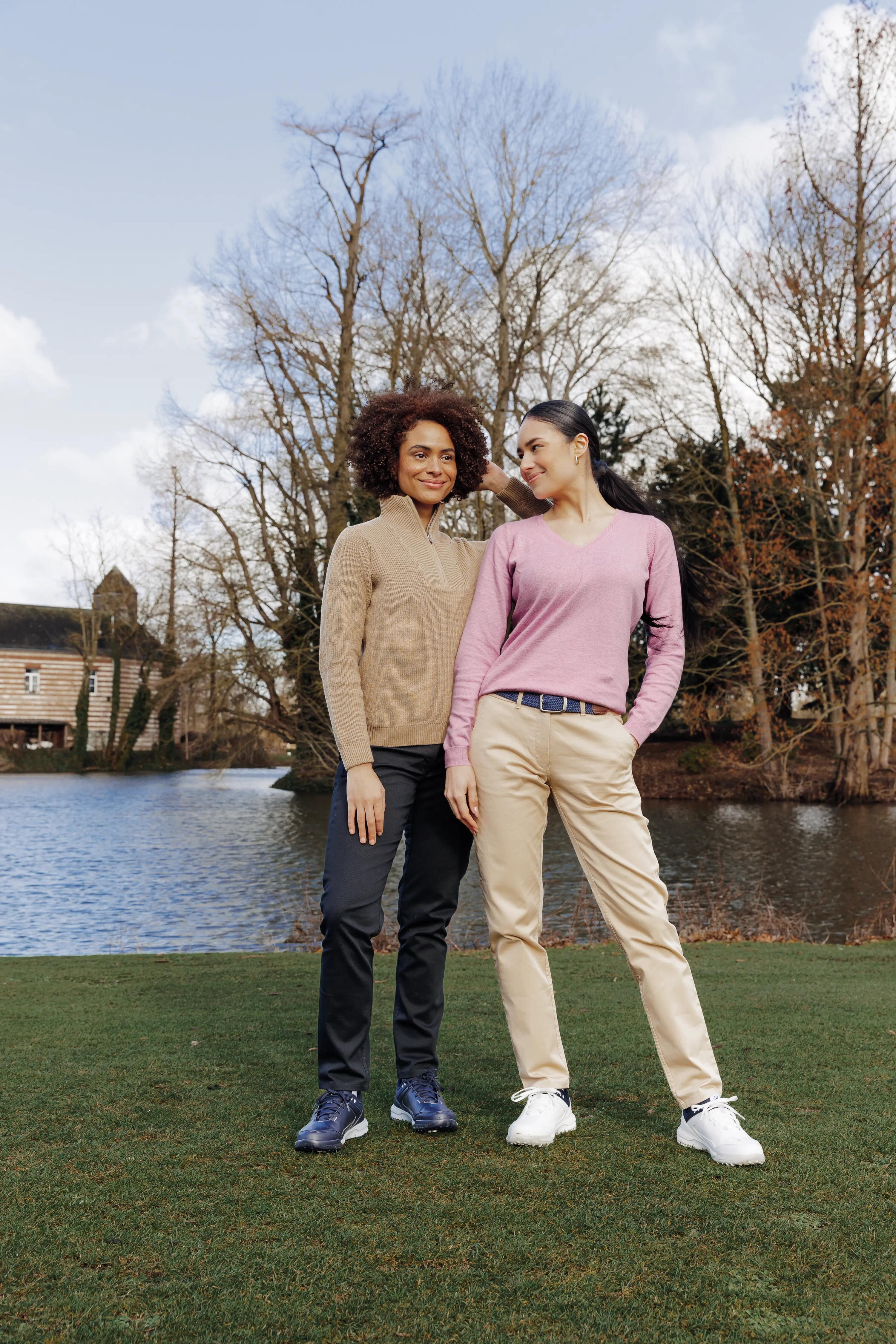 Zwei Frauen in stilvoller Golfkleidung stehen lächelnd auf einer Wiese vor einem See.