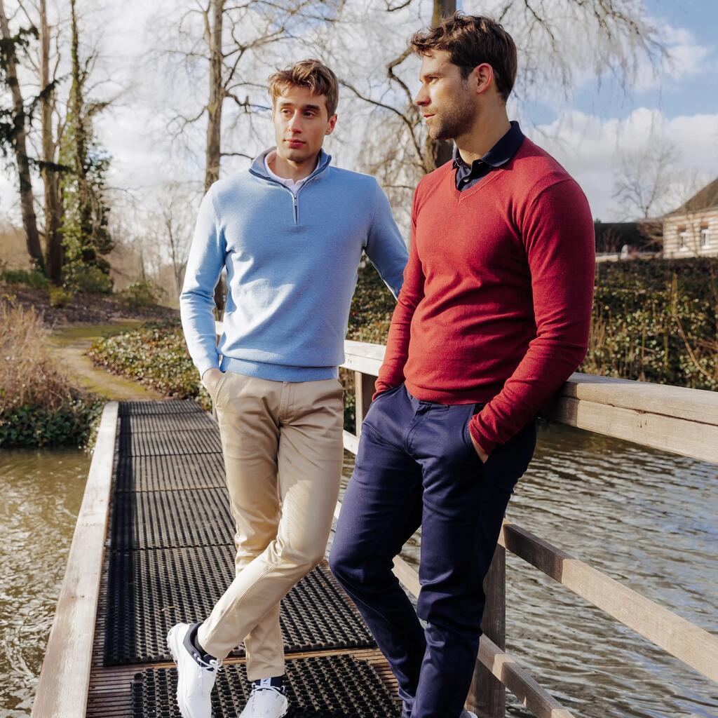 Zwei Männer in eleganter Golfbekleidung lehnen an einer Holzbrücke über einem Wasserlauf.