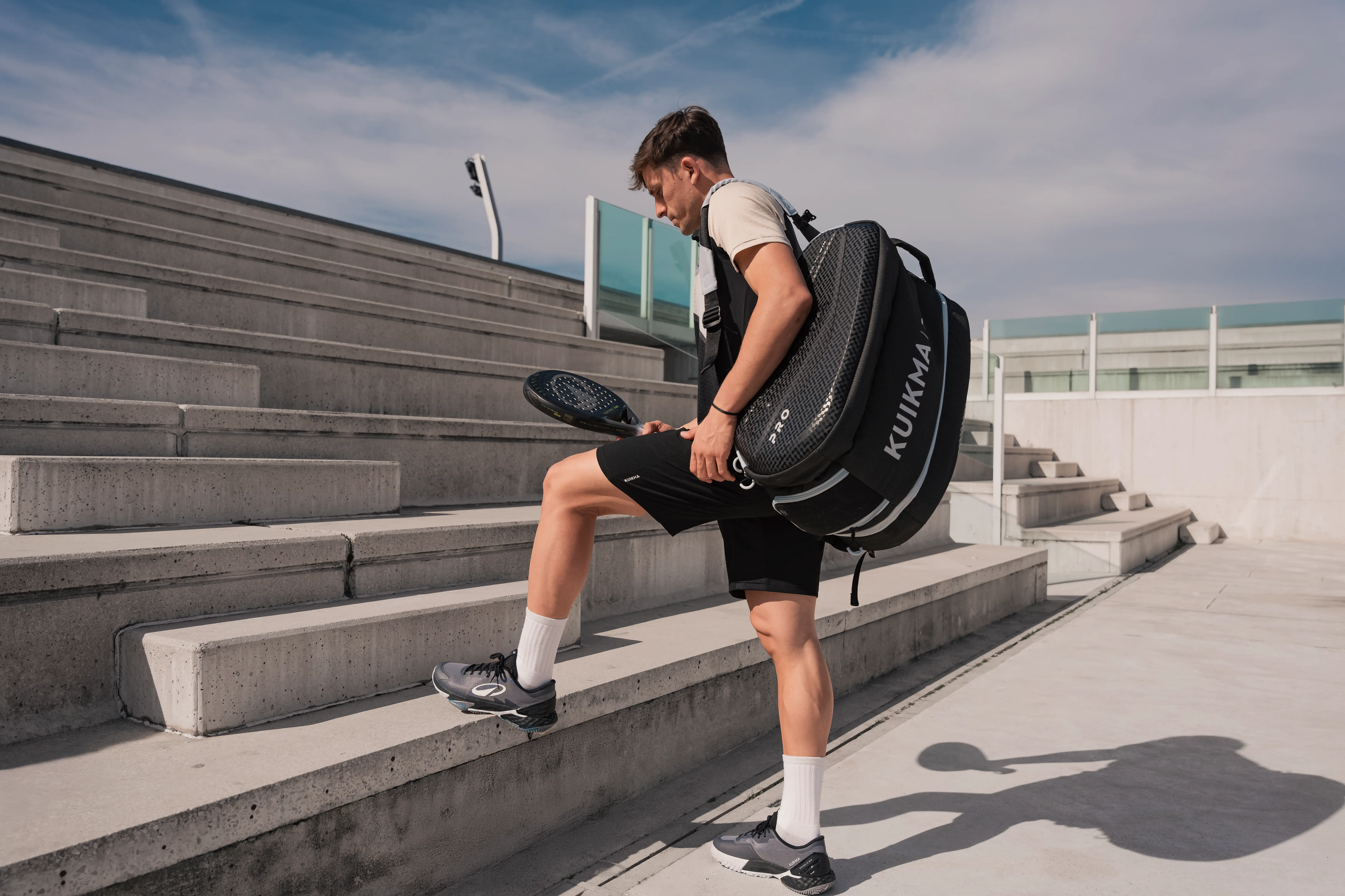 Ein Mann geht mit einer Kuikma-Padel-Tasche auf dem Rücken und einem Padelschläger in der Hand eine Betontreppe hinauf.