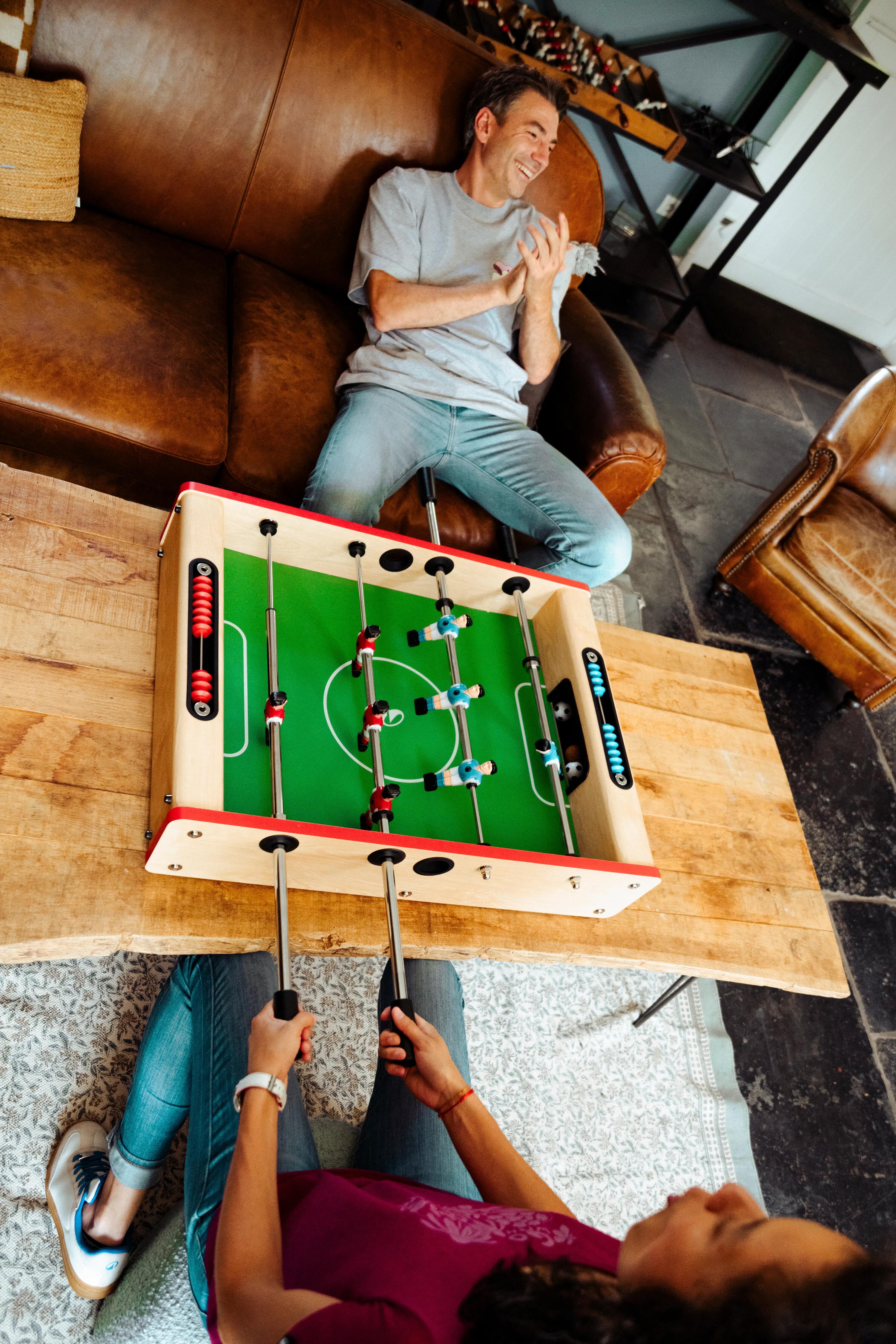 Ein Mann und eine Frau spielen lachend an einem kompakten Tischkicker aus Holz auf dem Couchtisch.