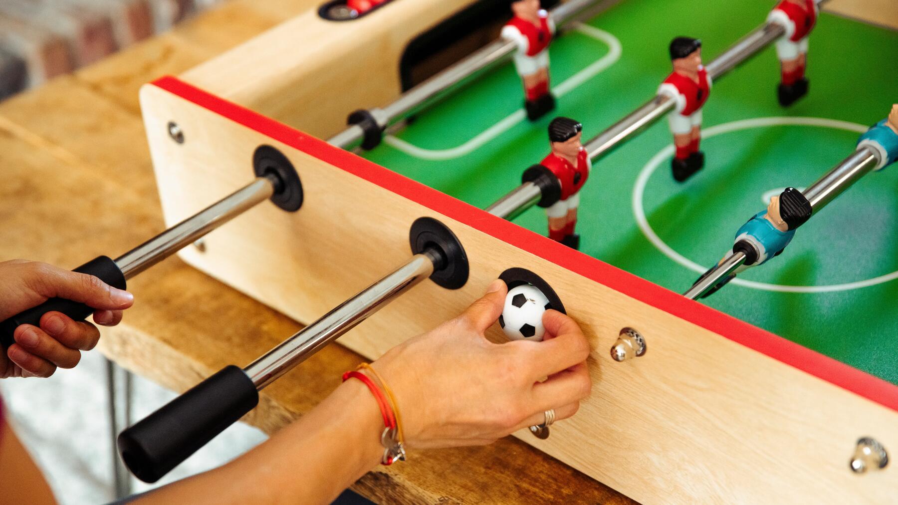 Eine Hand, die einen Mini-Fußball hält, neben einem hölzernen Tischkicker.