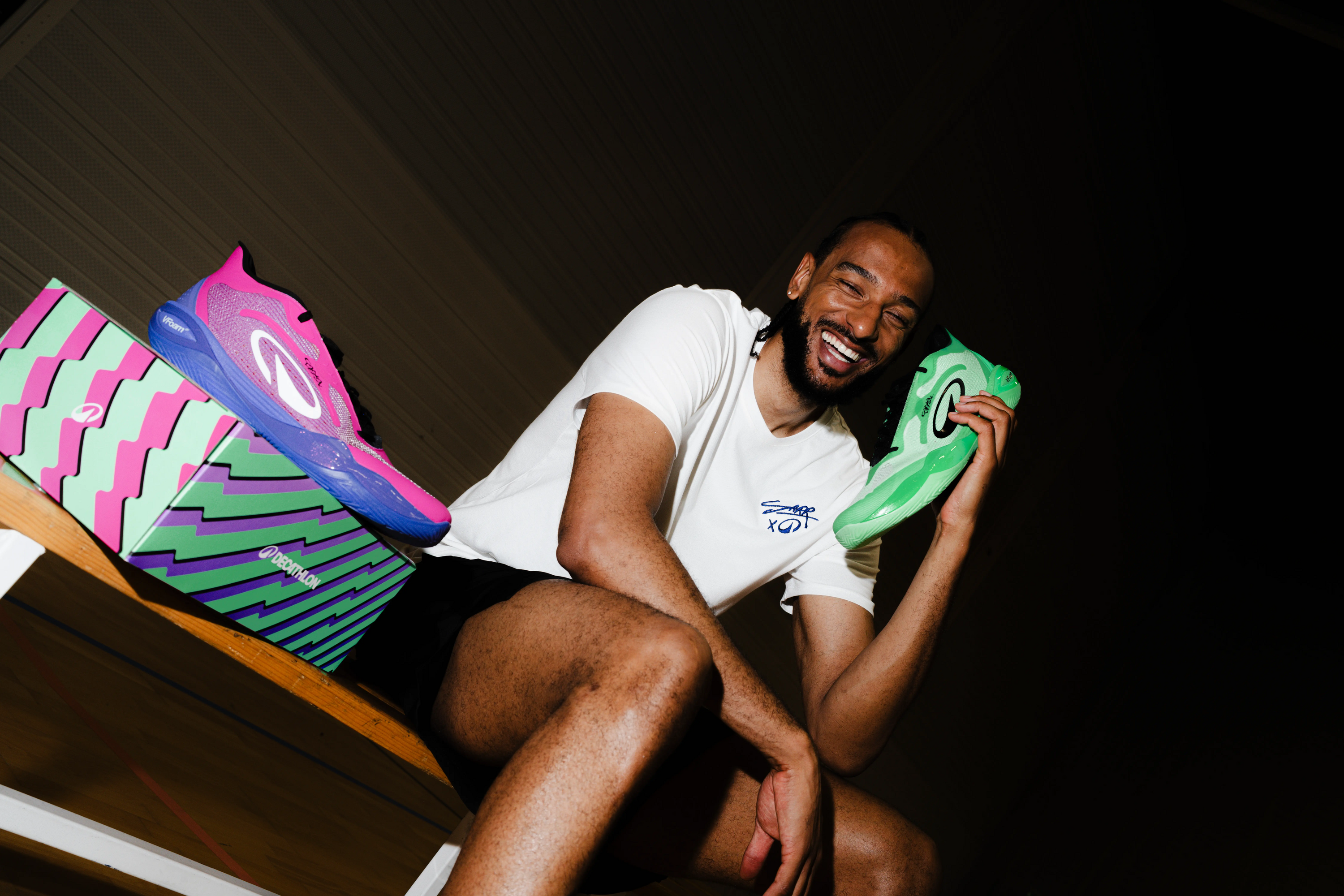 Ein lächelnder Mann sitzt mit einem grünen Sportschuh in der Hand neben einem farbenfrohen Schuhkarton und einem rosa-blauen Sportschuh.