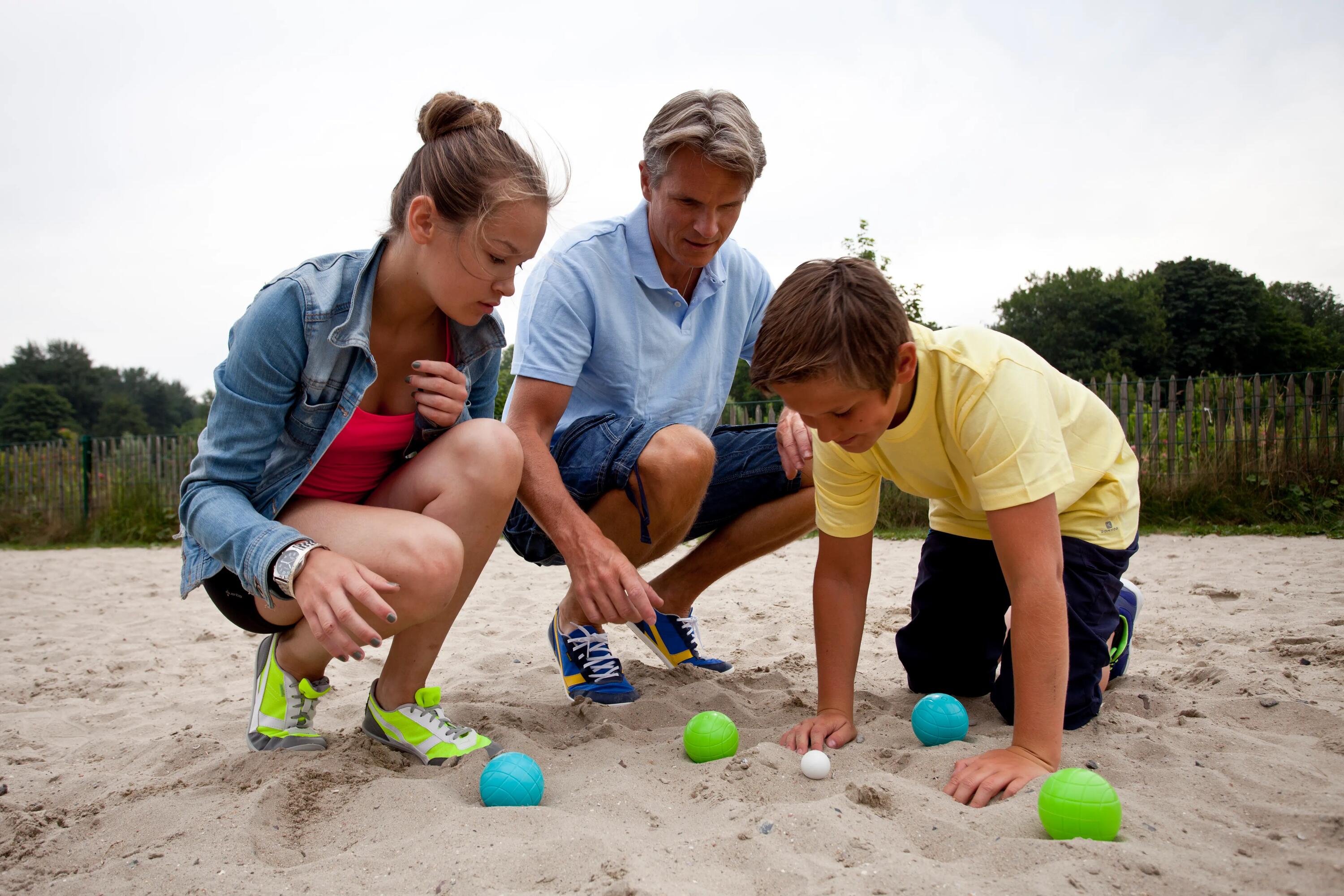 Eine Familie kniet im Sand und begutachtet die Lage bunter Boccia-Kugeln um die kleine weiße Zielkugel.