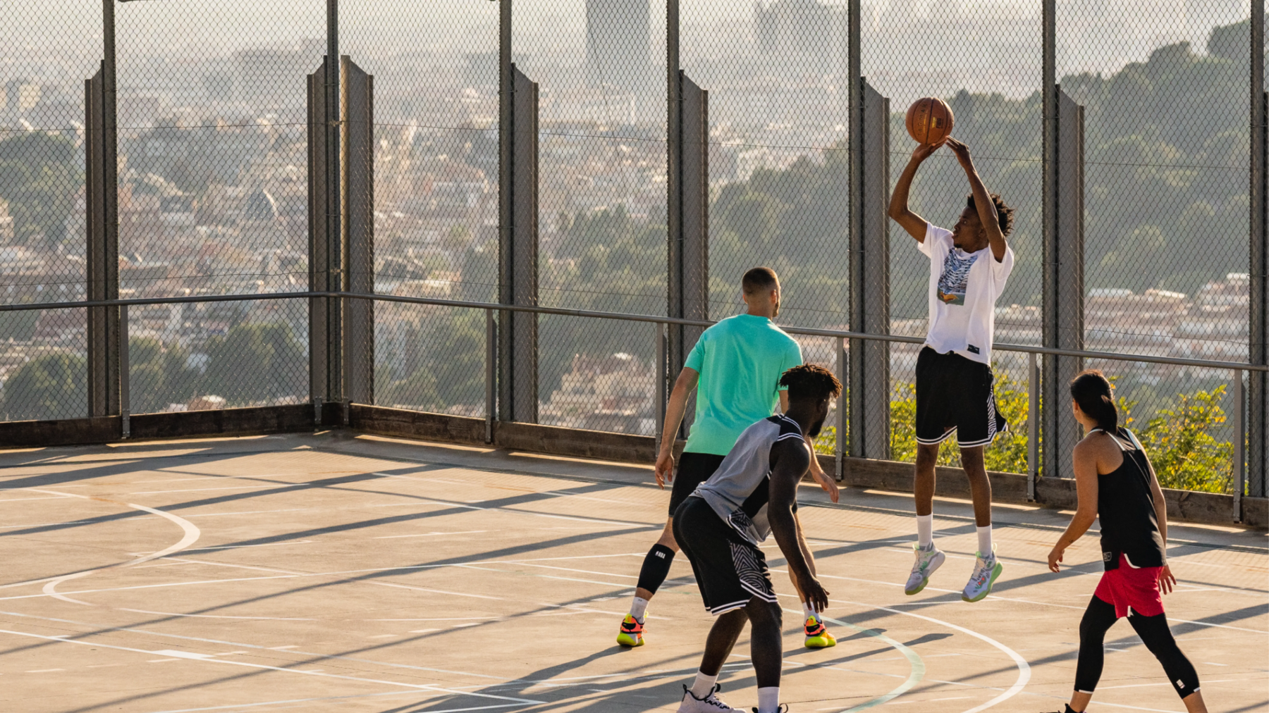 Drei Männer und eine Frau spielen Street Basketball auf einem Hügel. Stadt im Hintergrund.