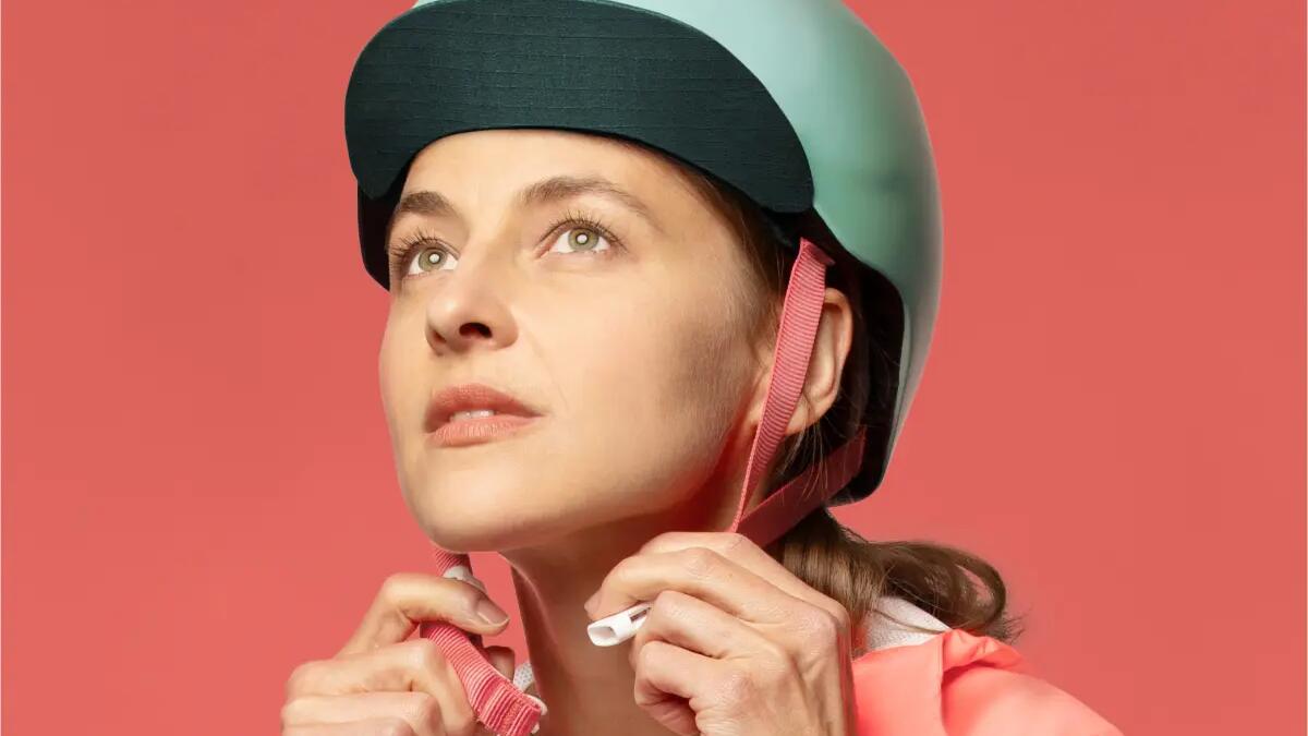 Frau mit hellgrünem Helm und rosa Jacke vor rosa Hintergrund, fokussiert nach oben schauend.