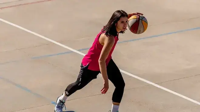 Frau im pinken Oberteil dribbelt bunten Basketball auf hellbraunem Betonplatz mit weißen Linien.