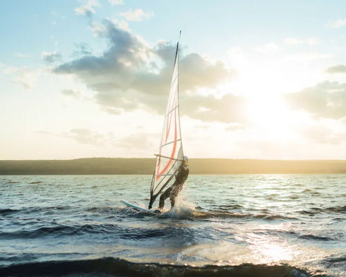 osoba płynąca na desce windsurfingowej trzymając żagiel windsurfingowy