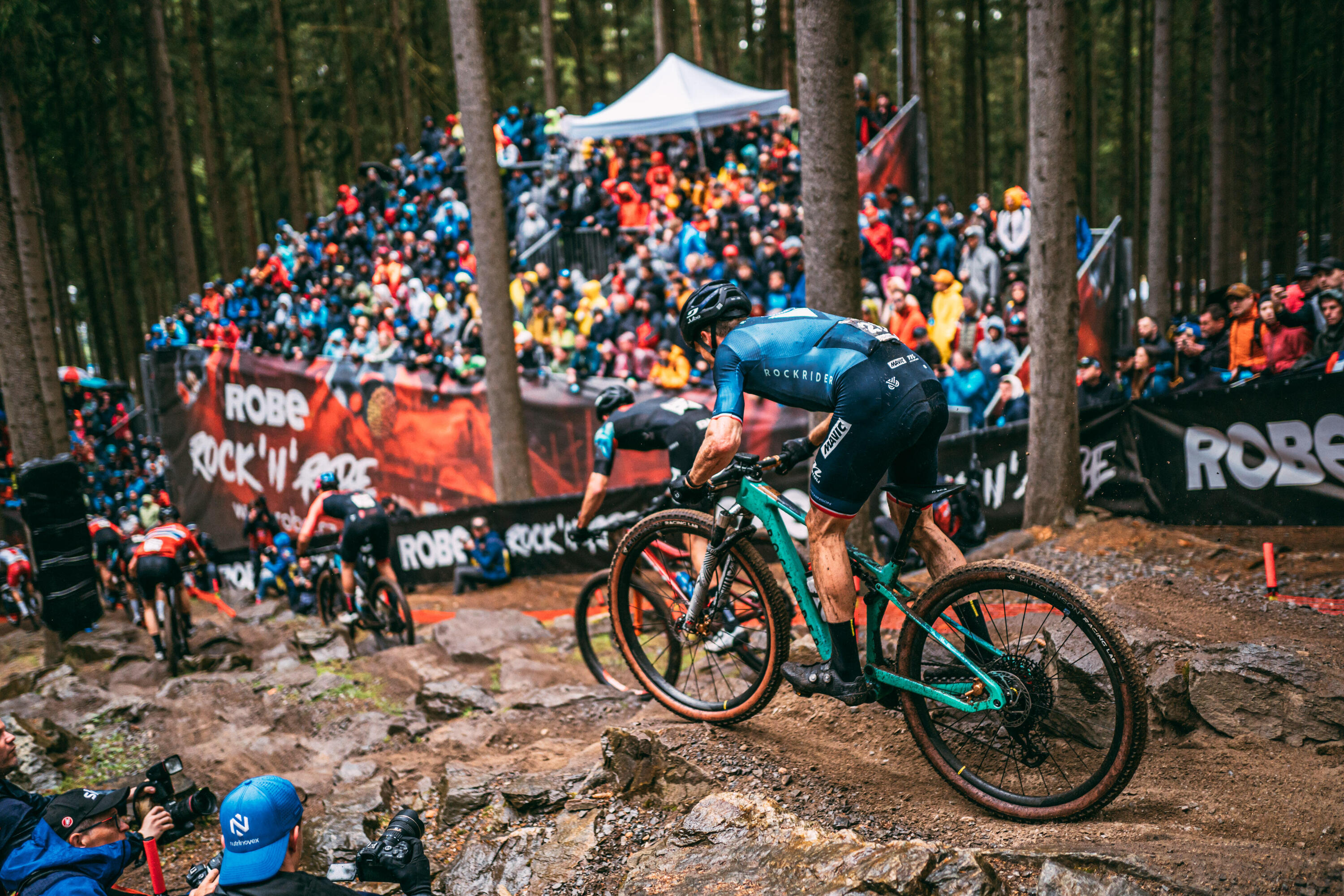 Ein Mountainbiker in blauer Rockrider-Teamkleidung fährt ein türkisfarbenes Mountainbike über einen steilen, felsigen Waldabschnitt während eines Rennens. Im Hintergrund säumen zahlreiche Zuschauer den Streckenrand im dichten Wald, während Fotografen im Vordergrund das Geschehen festhalten.