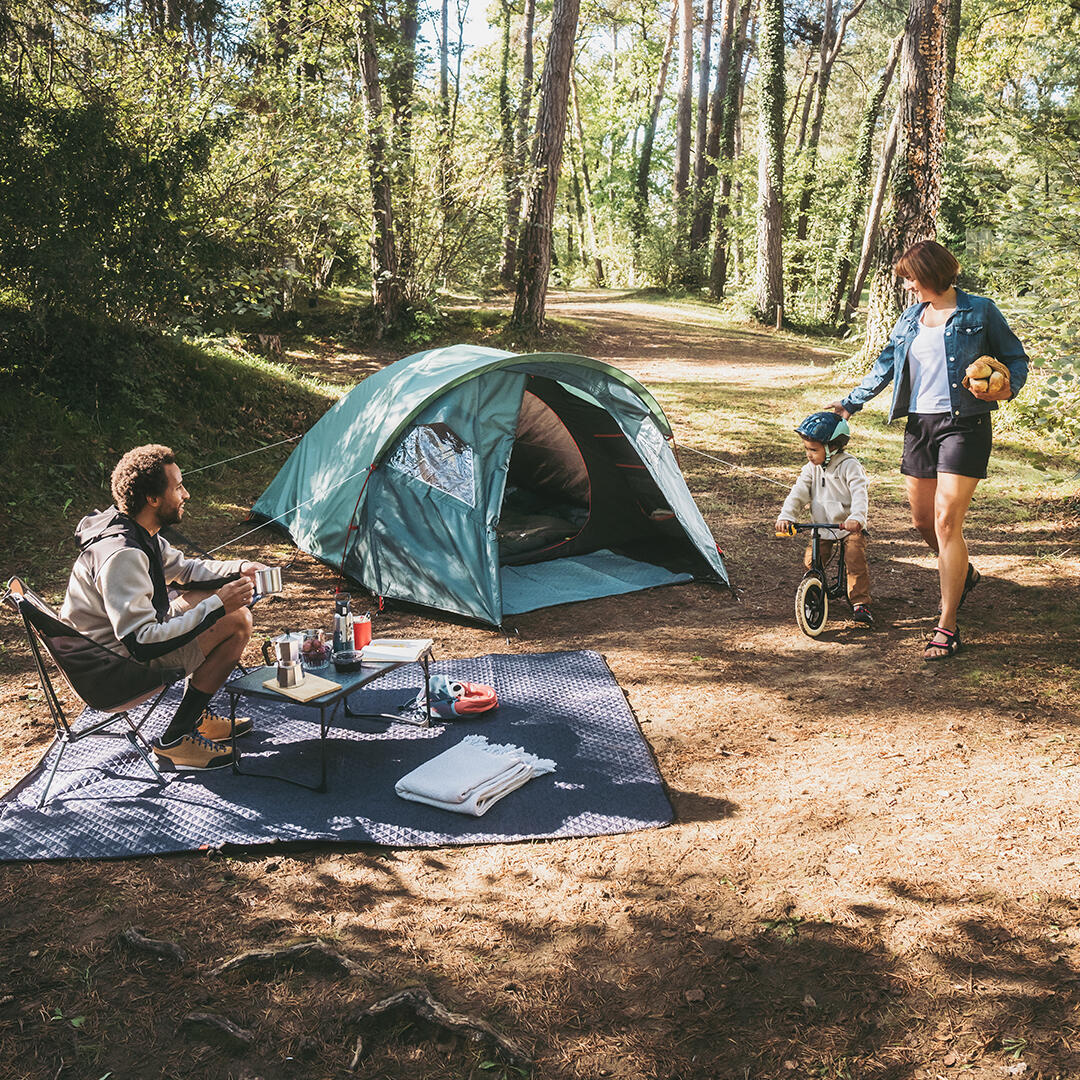 Zelt im Wald, grün, umgeben von Bäumen. Picknickdecke blauer Stoff, Waldweg, sommerliche Atmosphäre. Eltern mit Kind, Fahrrad.