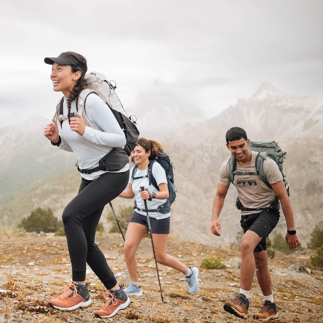 Drei wandernde Personen in sportlicher Kleidung, fröhlich auf einem Bergpfad. Grauer Himmel, steiniger Boden, grüne Bäume am Horizont.