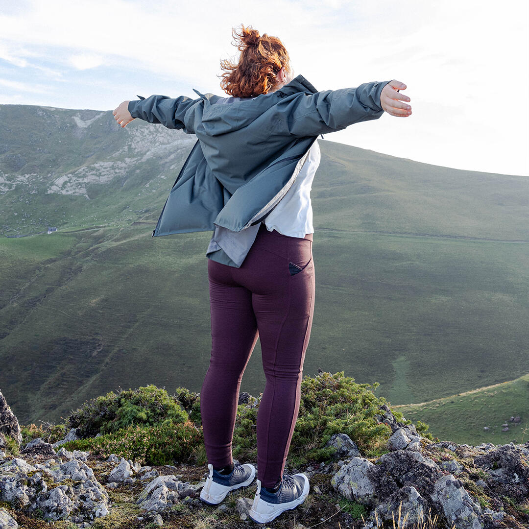 Person mit ausgebreiteten Armen auf hügeliger Landschaft, blauer Himmel, tiefgrüne Hügel und Steintexturen.