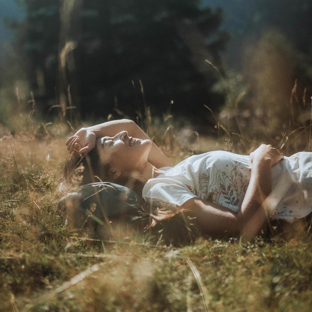 Frau liegt lächelnd im Gras, Sonnenstrahlen, helle Farben, weiches Licht, entspannte Szene in natürlicher Umgebung.