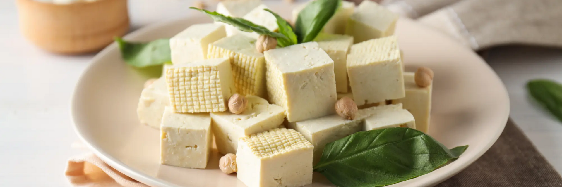 Grafika przedstawiająca talerz z produktem wegańskim pokrojonym w kosteczkę tofu ozdobionym liściami bazylii i orzechami laskowymi