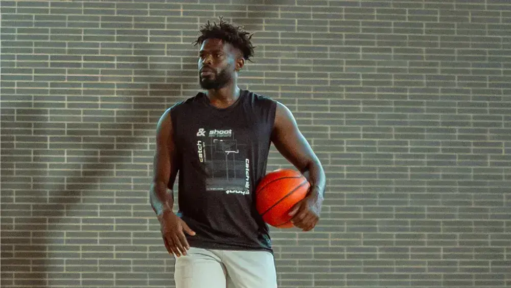 Mann mit schwarzem Shirt hält orangen Basketball; graue Backsteinwand im Hintergrund; lässige Pose, sportlicher Stil.