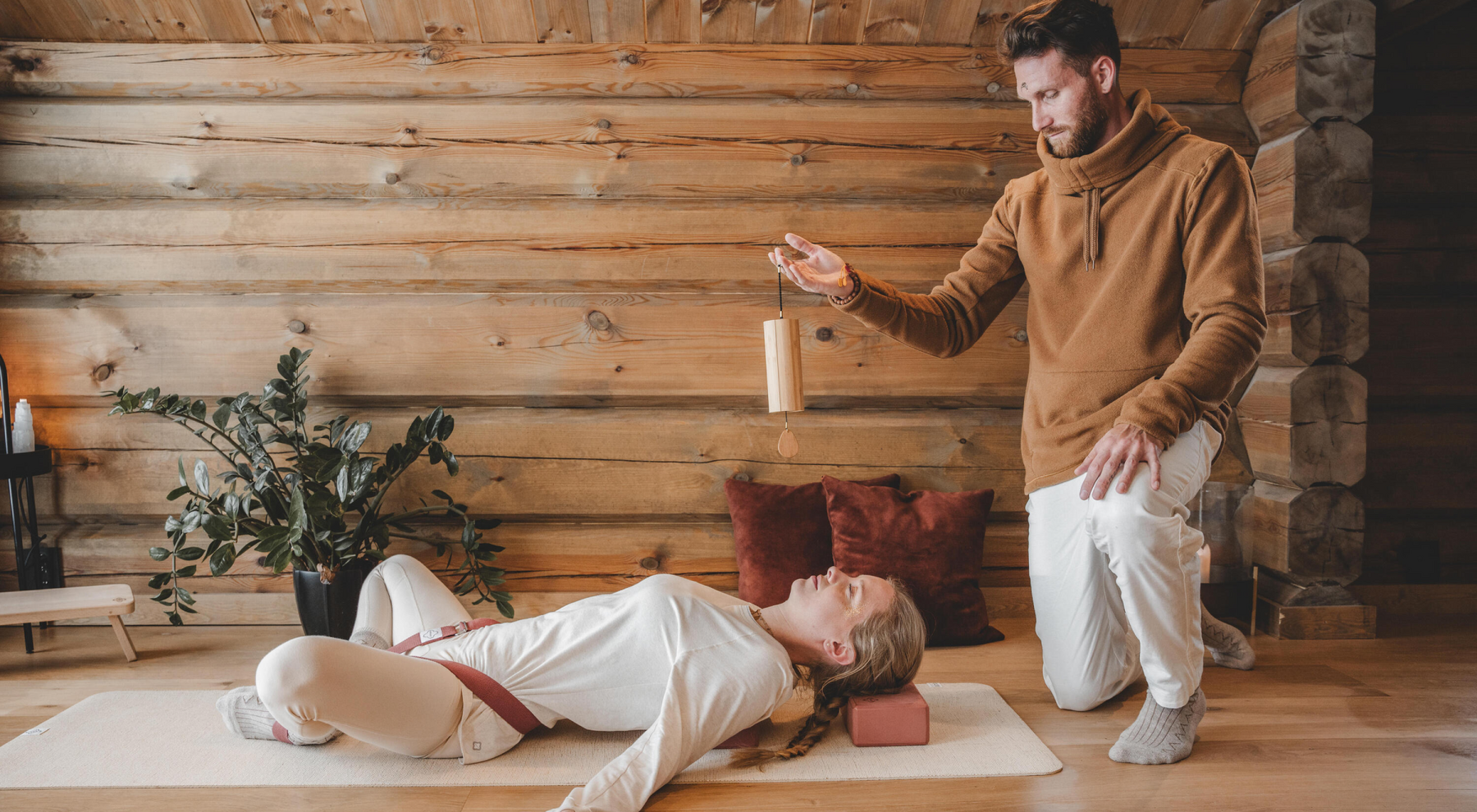 Kobieta leżąca w pozycji supta baddha konasana na macie nad nią kuca instruktor i macha kadzidłami