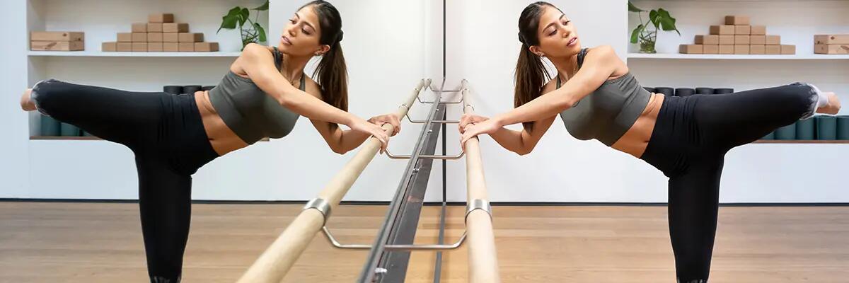 Kobieta w stroju sportowym ćwicząca barre pilates przed lustrem trzymając się drążka