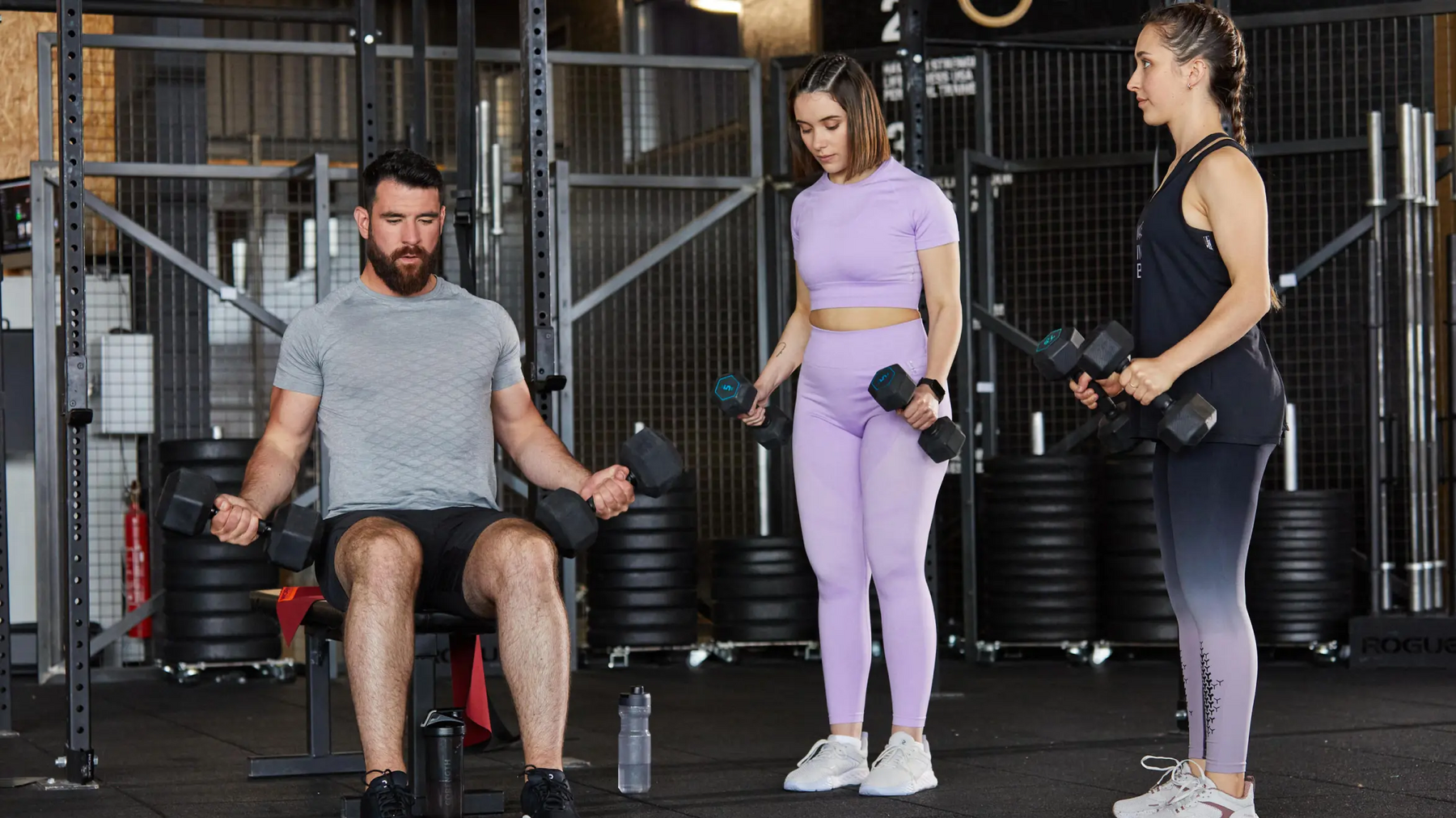 Mężczyzna siedzący na ławce ćwiczący arnoldki obok dwie kobiety ćwiczące arnoldki na stojąco