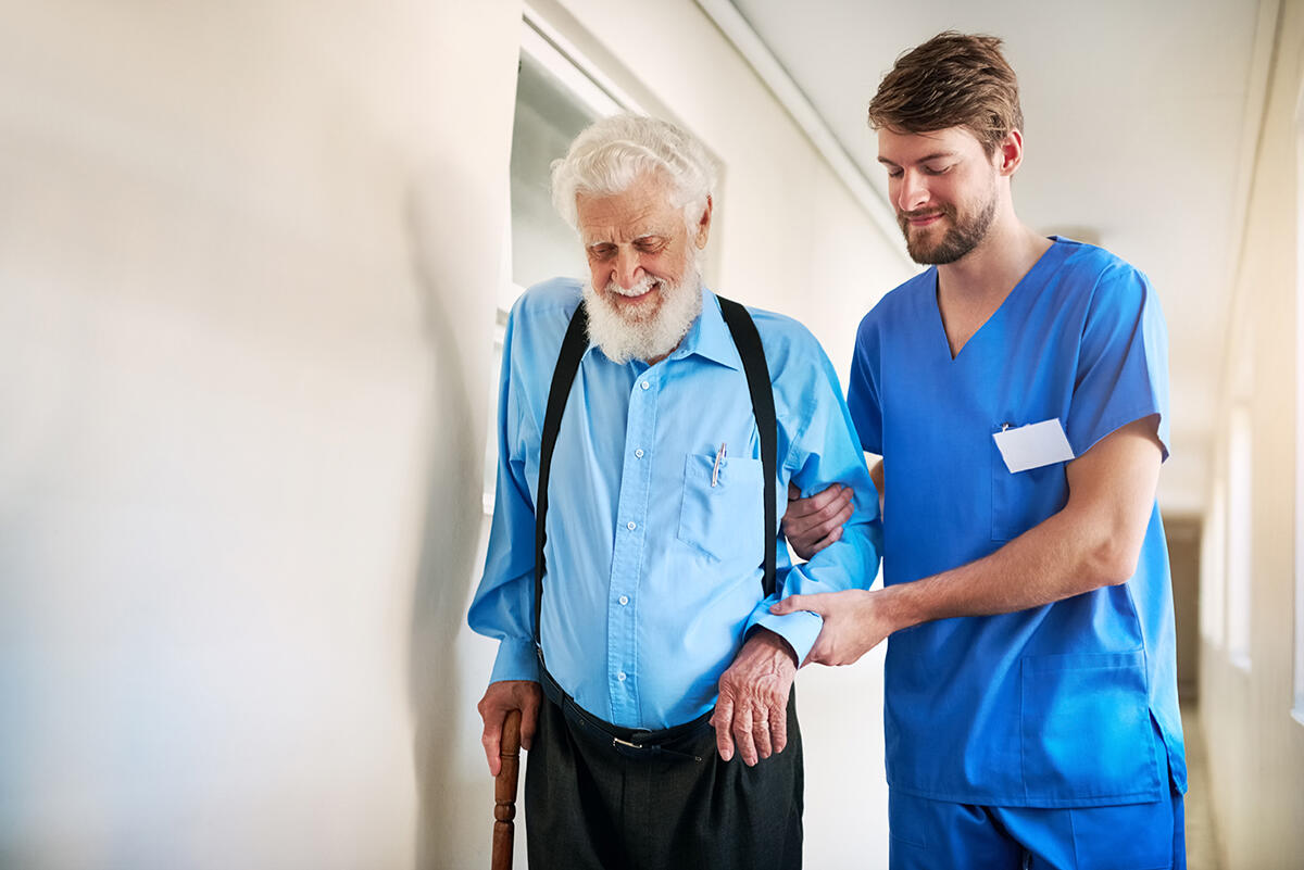 Rehabilitant podtrzymujący seniora za rękę idącego o lasce