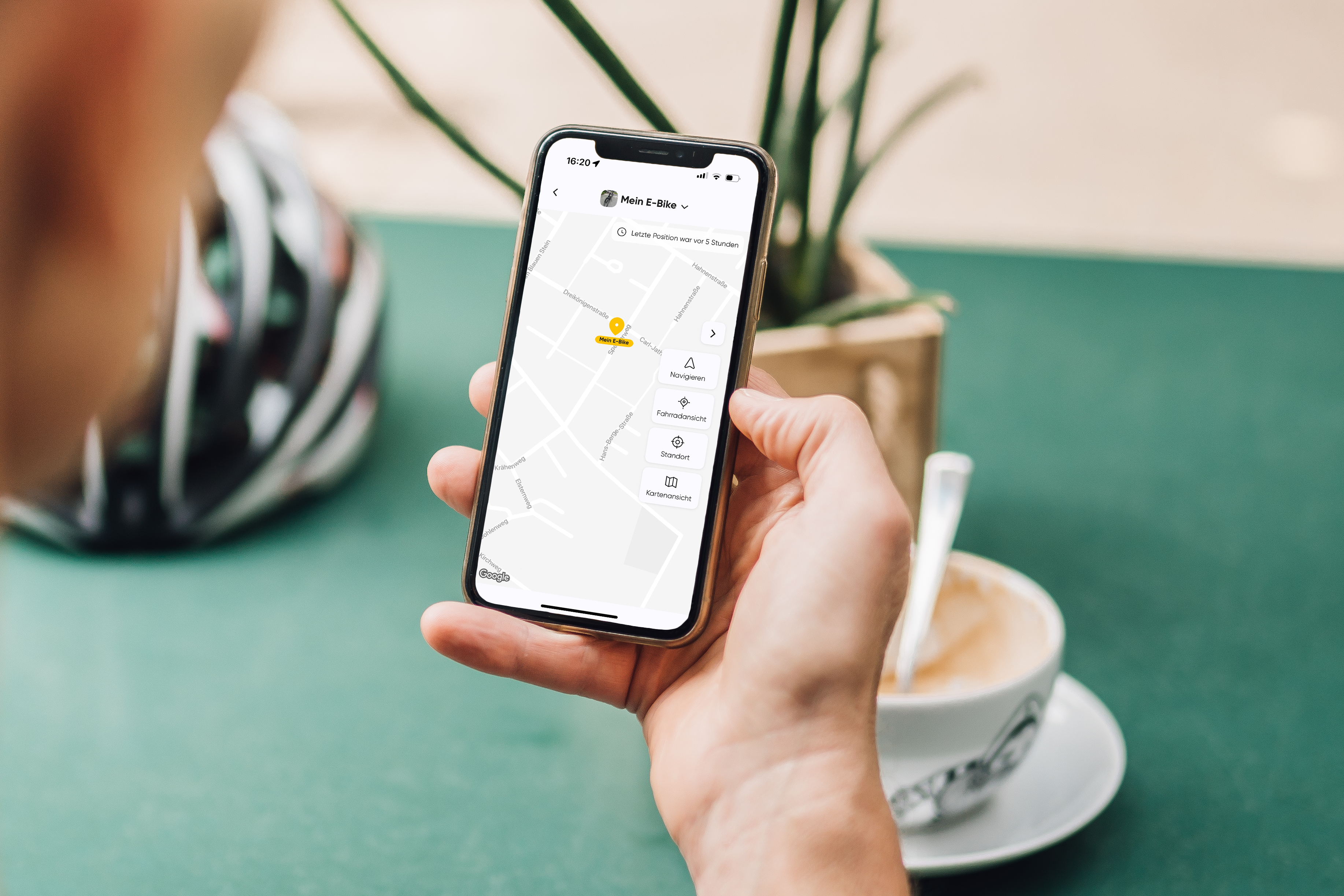 Smartphone in Hand mit Karten-App, grüner Tisch, Kaffeetasse, Fahrradhelm, Pflanze im Hintergrund.