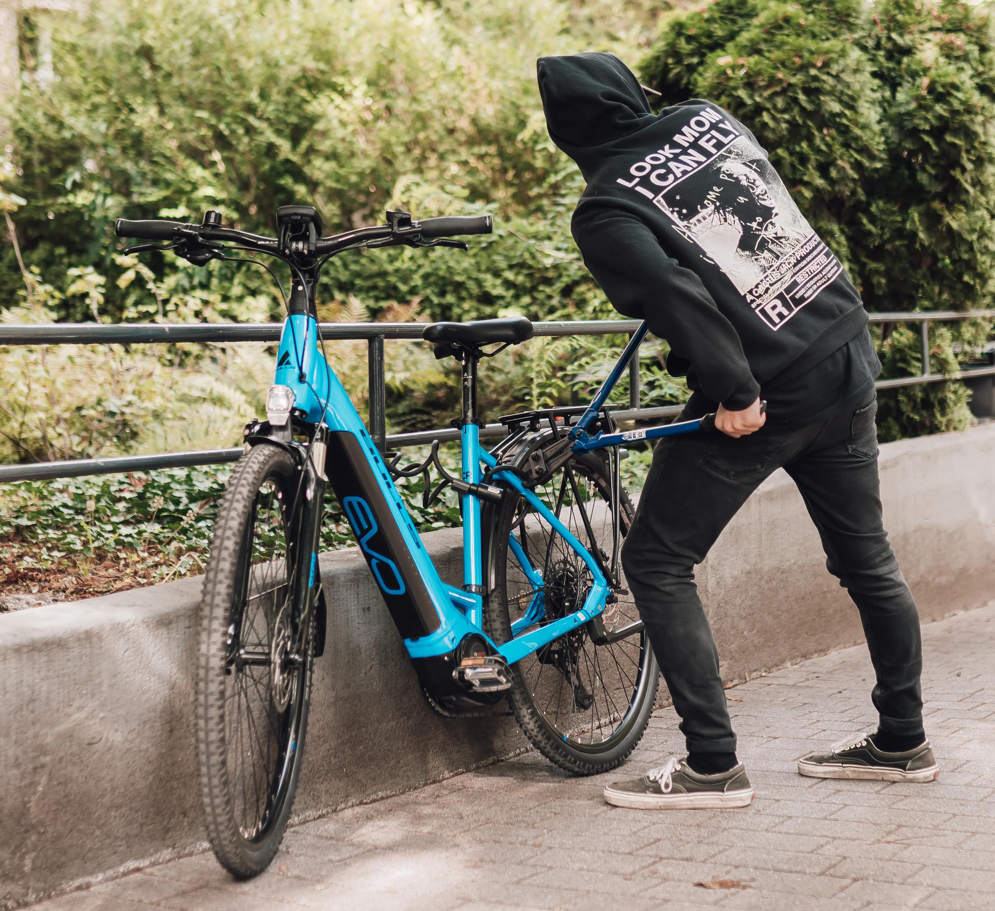 Person in schwarzem Hoodie stiehlt blaues Fahrrad; grüne Büsche im Hintergrund; Radweg aus Pflastersteinen.