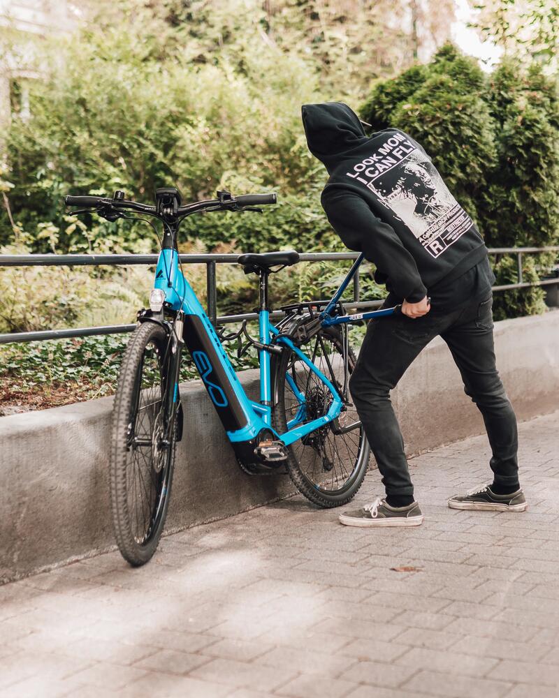Person schließt blaues Fahrrad an Geländer an, schwarzer Hoodie und Jeans, Kopf nach unten, grüne Büsche im Hintergrund.