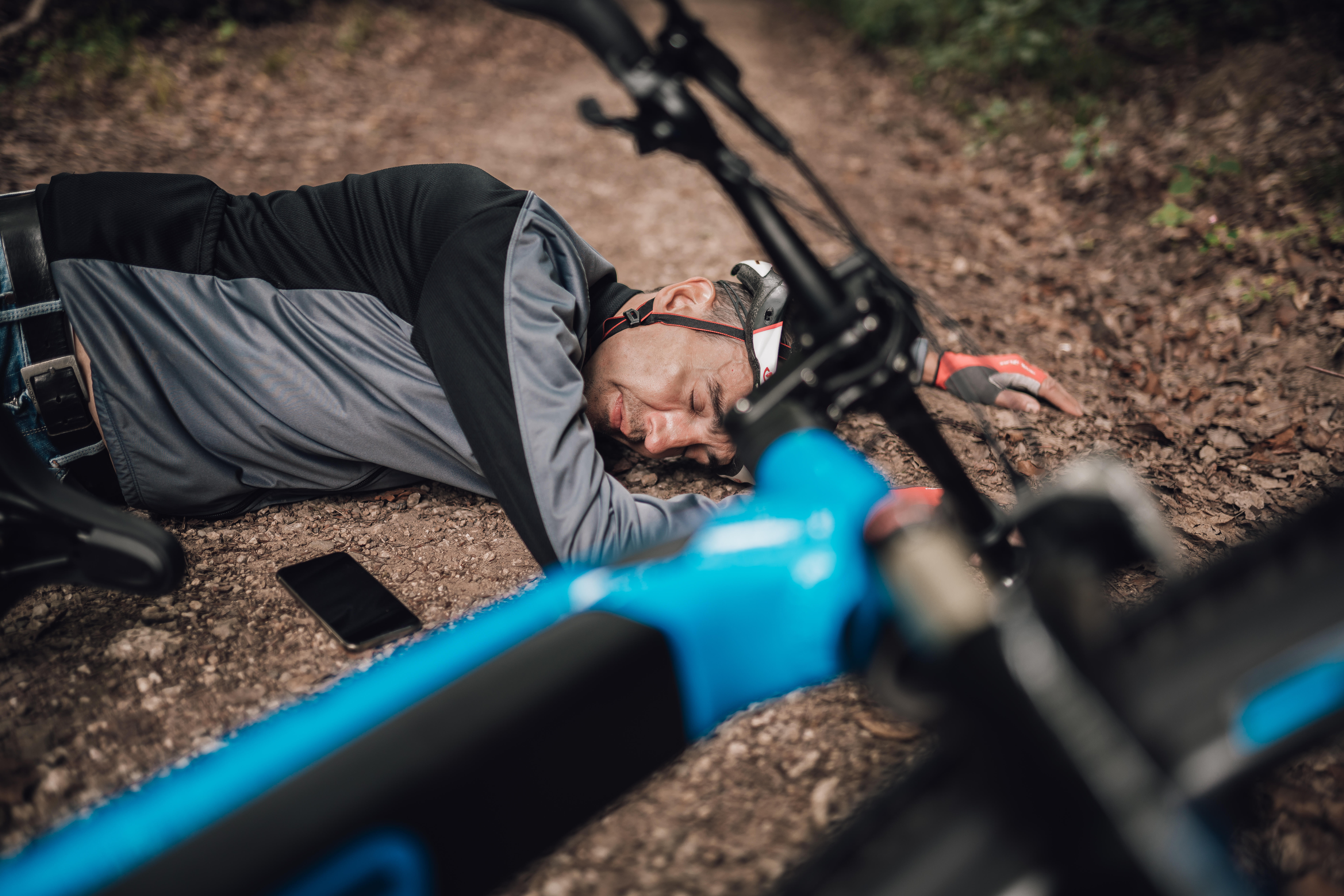 Person liegt auf Waldboden neben Fahrrad und Telefon. Blaue und schwarze Farben dominieren. Erschöpfter Ausdruck.
