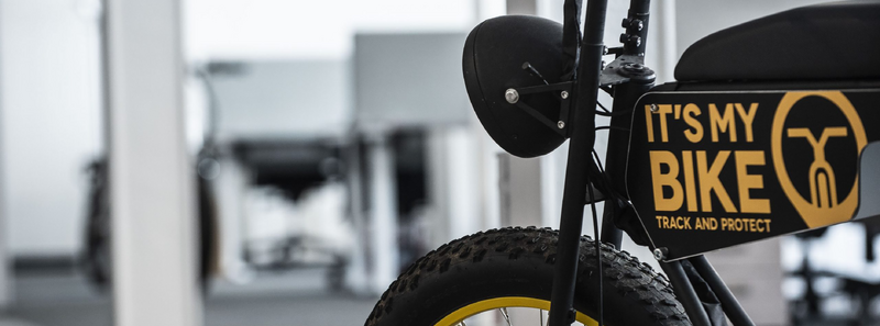 Schwarzes Fahrrad, gelbe Felgen, markanter Schriftzug, glatte Textur, industrielles Umfeld, dezente Grautöne im Hintergrund.