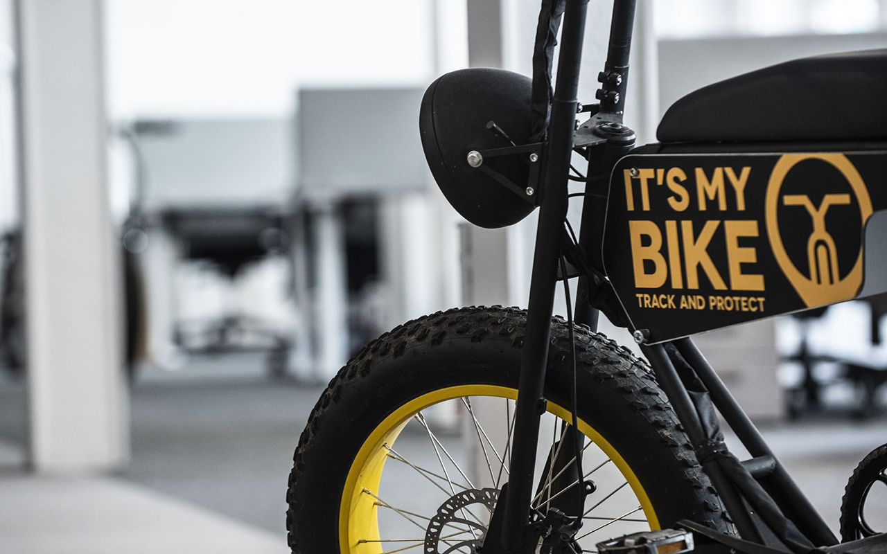 Schwarzes Fahrrad, gelbe Felgen, markanter Schriftzug, glatte Textur, industrielles Umfeld, dezente Grautöne im Hintergrund.