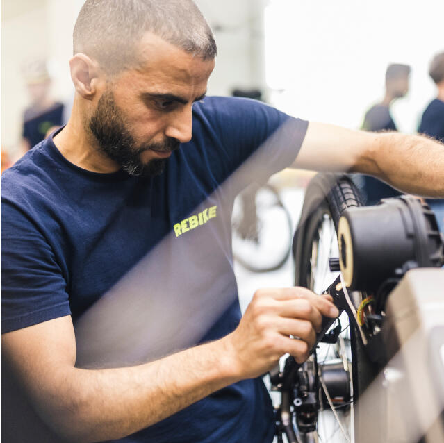 Ein Fahrradmechaniker im Rebike-T-Shirt repariert konzentriert ein Fahrrad in einer Werkstatt.