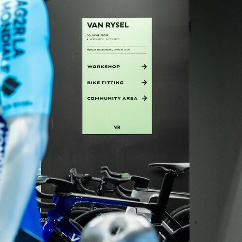 Blaues Fahrradtrikot, schwarze Fahrräder, weiß-grünes Schild mit schwarzem Text, schwarzer Hintergrund.