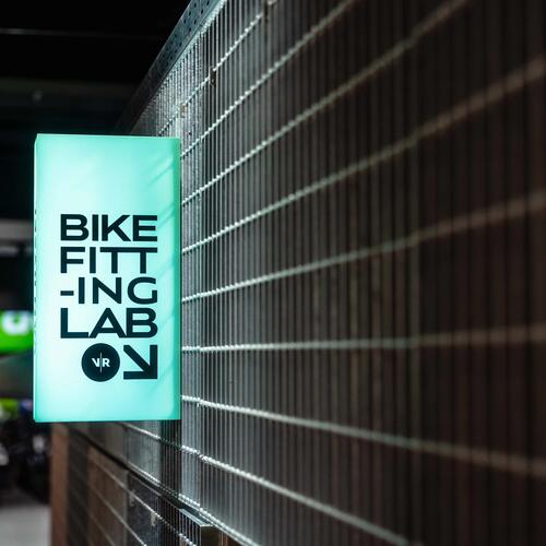 Grünes Leuchtschild mit schwarzem Text "BIKE FITTING LAB" an braun verkleideter Wand.