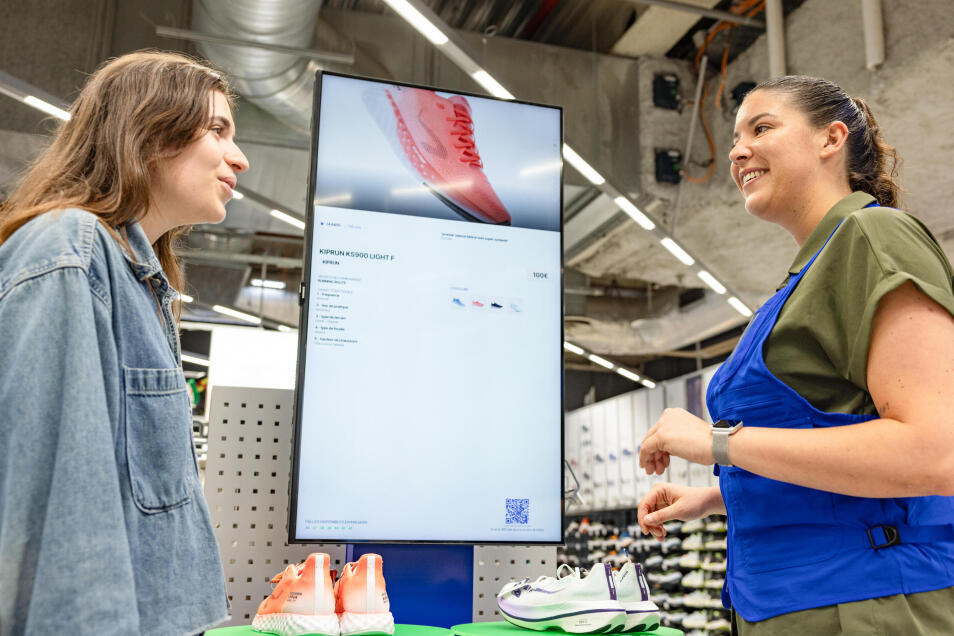 Eine Kundin lässt sich von einer Decathlon Mitarbeiterin beraten. Sie stehen vor einem Vertikalen Bildshirm, auf dem eine Produktdetailseite zu sehen ist. Vor dem Bildschirm auf einer Ausstellungsfläche stehen zwei Paare Laufschuhe.