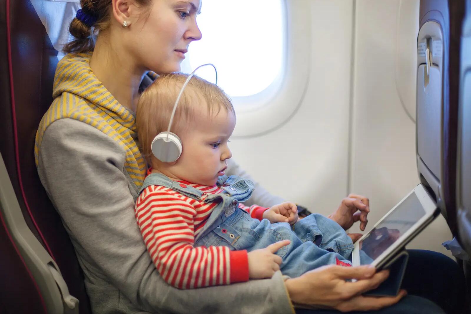Mama siedząca w samolocie trzymająca dziecko ze słuchawkami na uszach na kolanach pokazująca mu coś na tablecie