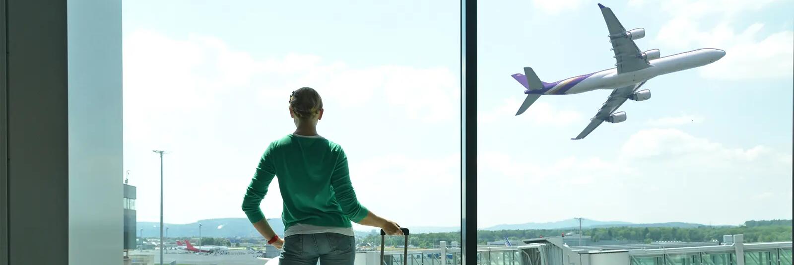 Kobieta z walizką podręczną patrząca przez okno na lotnisku jak startuje samolot korzystająca z porgramu lojalnościowego Miles&More
