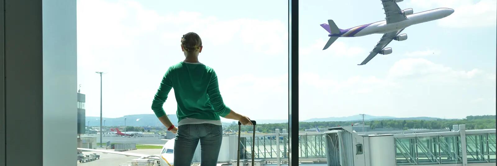 Kobieta trzymająca walizkę podręczną patrząca przez okno na lotnisku jak startuje samolot