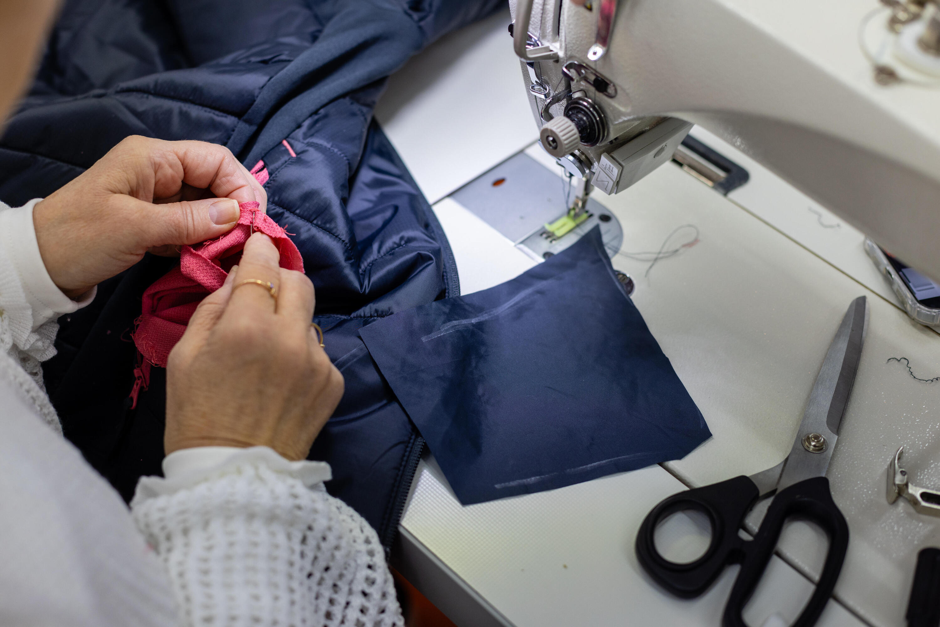 Eine Person die an einer Nähmaschine präzise mit Textilien (einer blauen Jacke und pinkem Stoff) arbeitet. Neben der Nähmaschine liegt eine Schere auf dem Arbeitstisch.