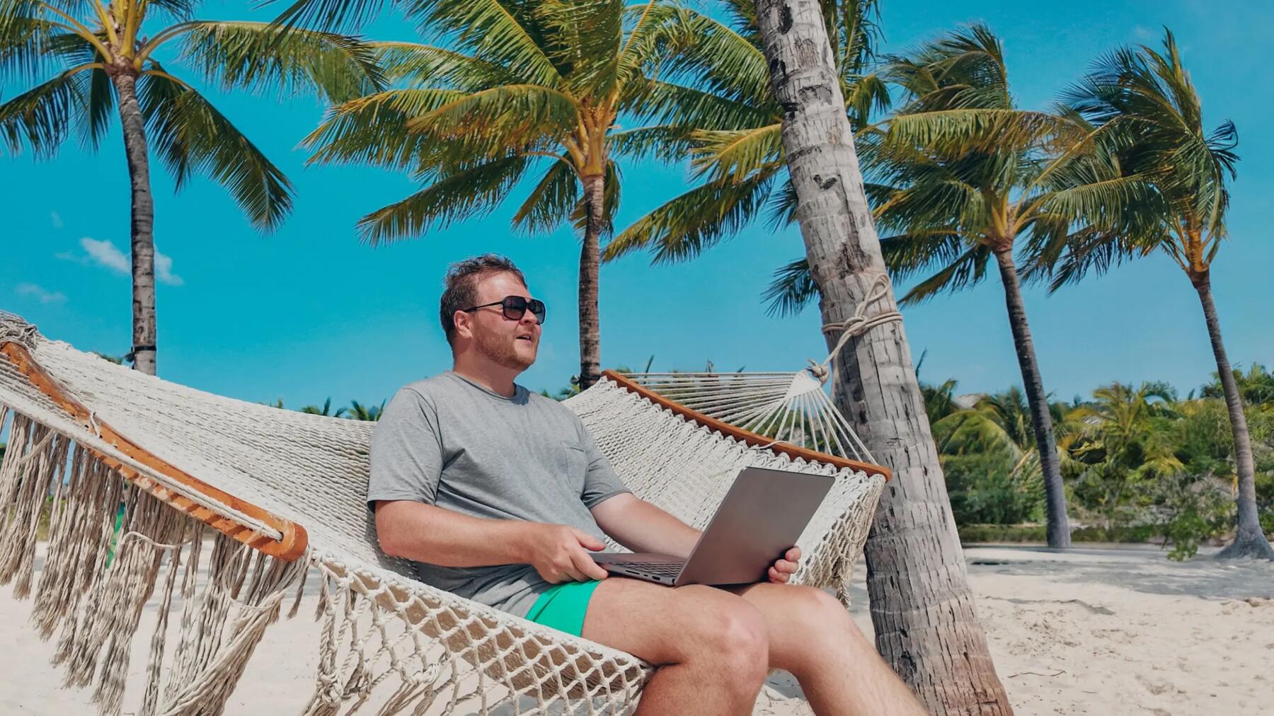Mężczyzna siedzący na hamaku wśród palm z laptopem na kolanach